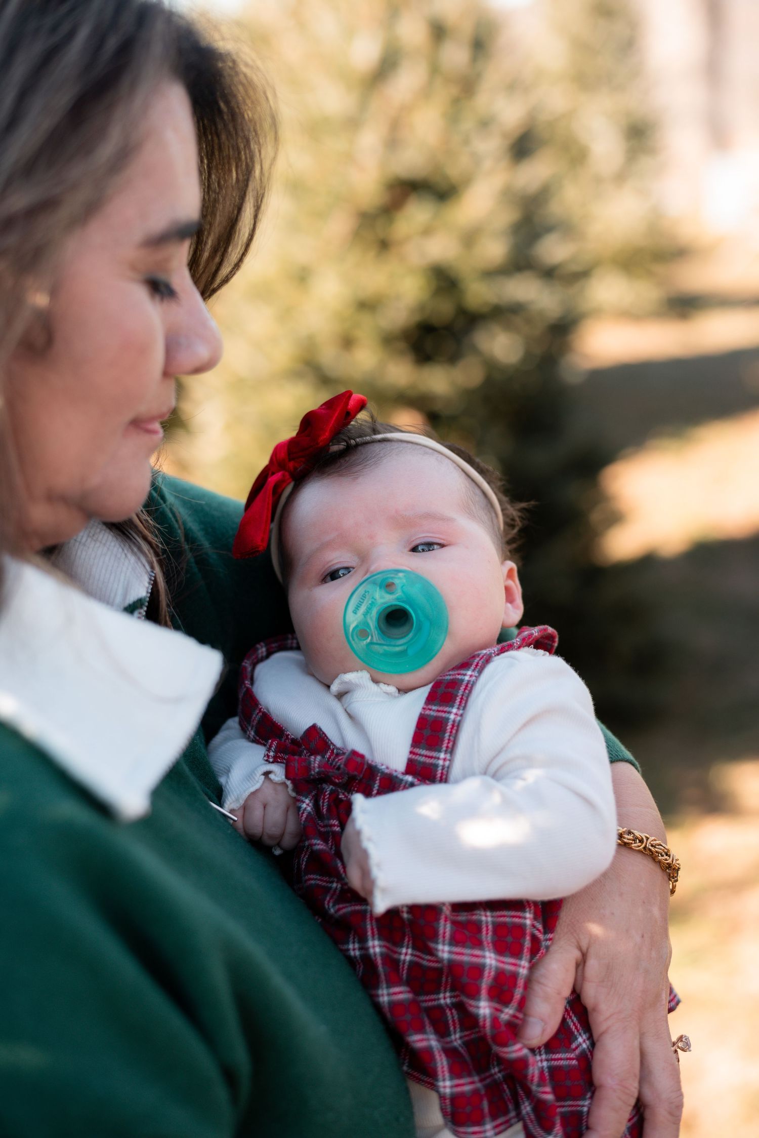 Parent in green sweater holding baby wearing plaid outfit and pacifier.