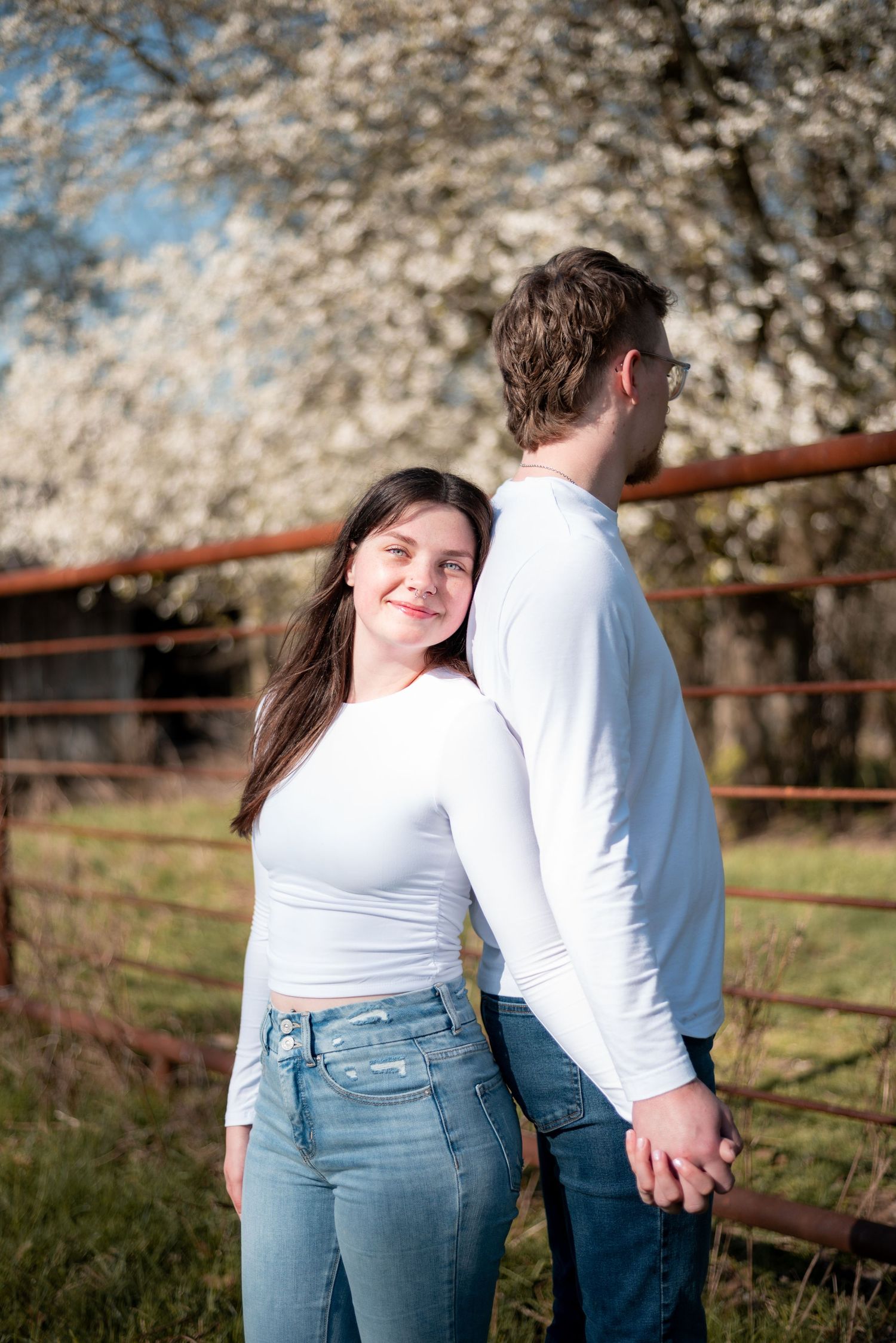 A young couple in matching white tops and jeans hold hands near a rustic fence with blooming white trees in spring.