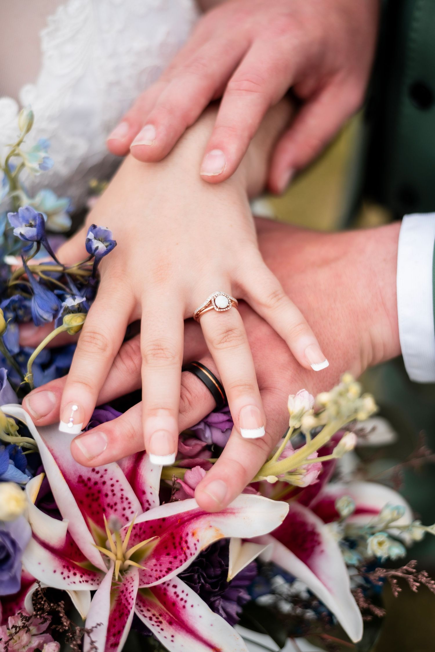 Newlyweds hands stacked on colorful wedding bouquet with stargazer lilies, bride wearing gold ring with French manicure.