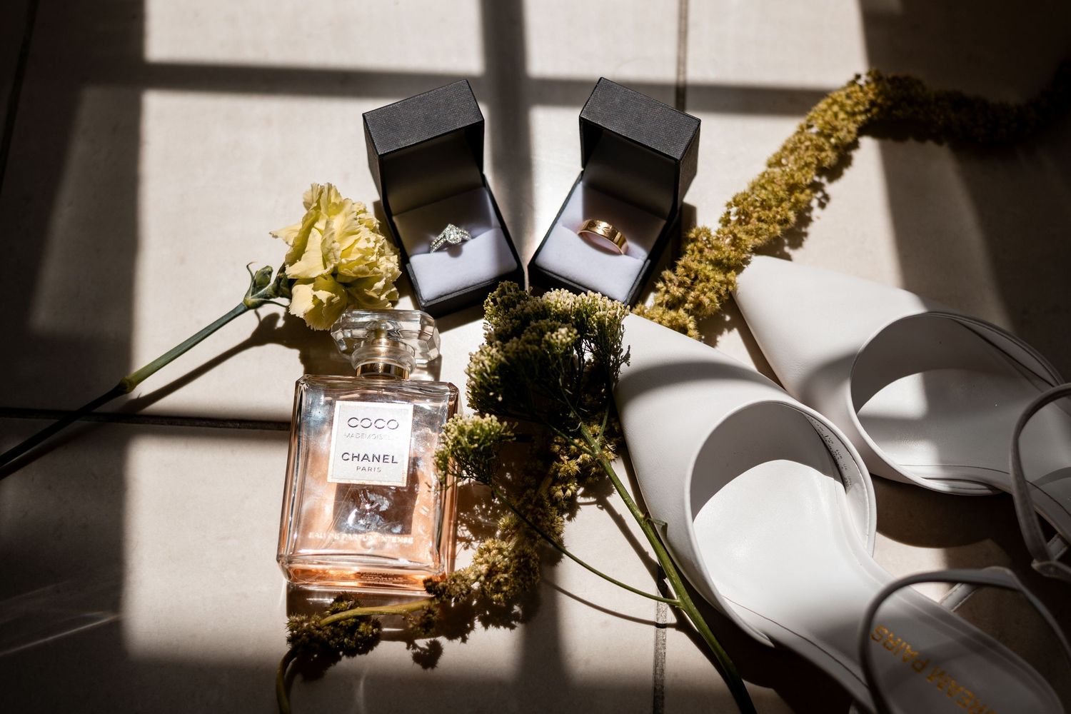 Chanel Coco perfume, wedding rings in boxes, white heels, and yellow flowers arranged in sunlight.