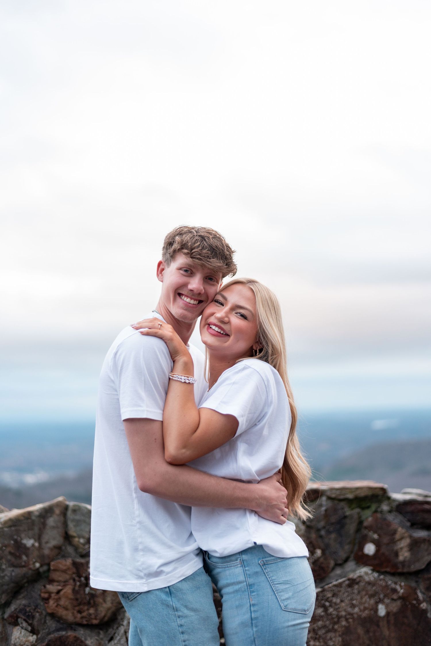 A young couple in matching white t-shirts and jeans embrace and smile on a rocky overlook with a cloudy sky backdrop.