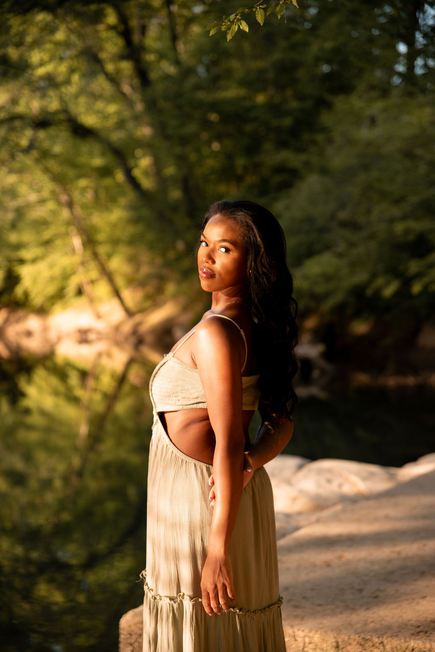 Young woman in sage green two-piece maxi outfit posing by a sunlit forest river on rocky terrain.