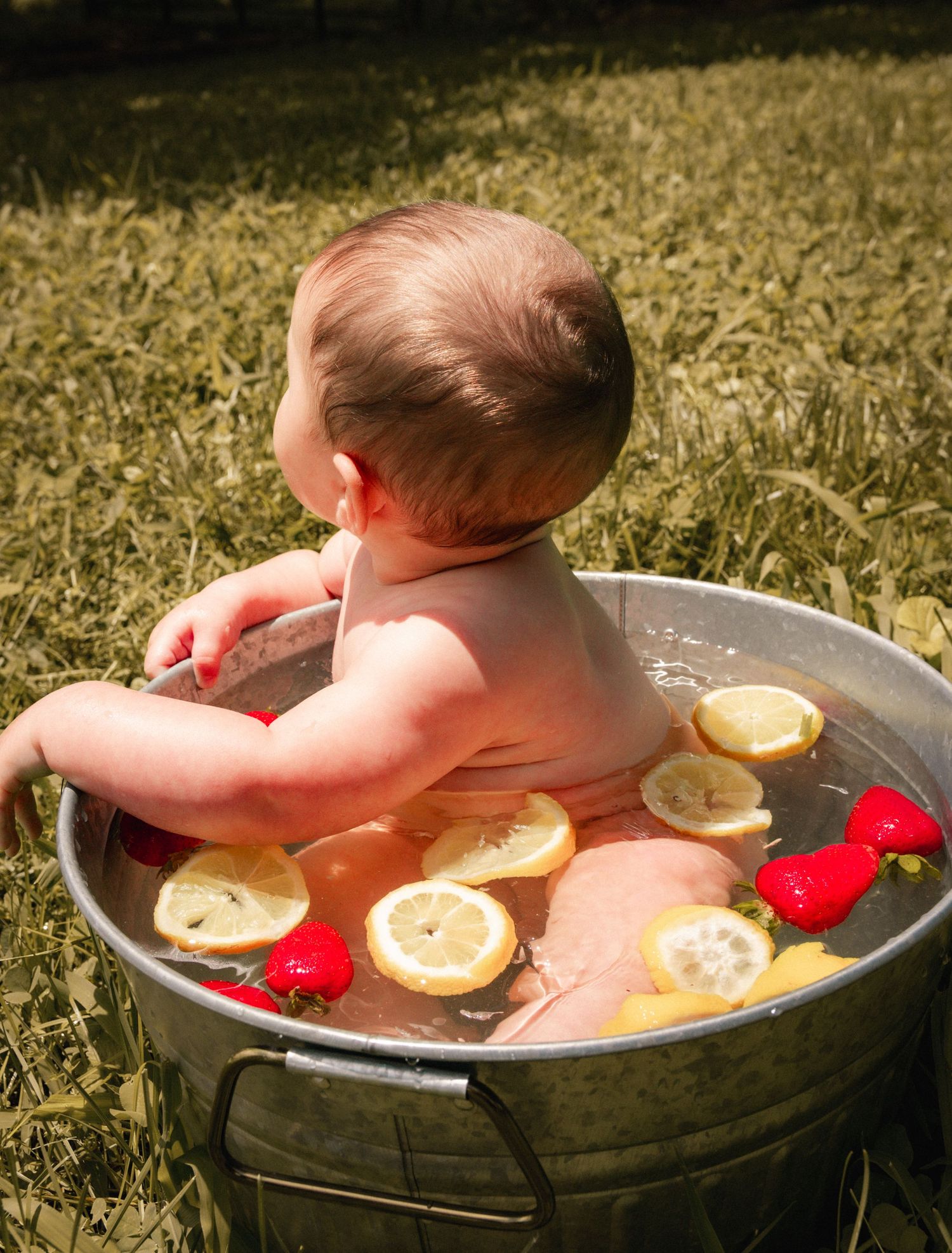 Baby taking a bath in metal tub with lemon slices and flowers outdoors.