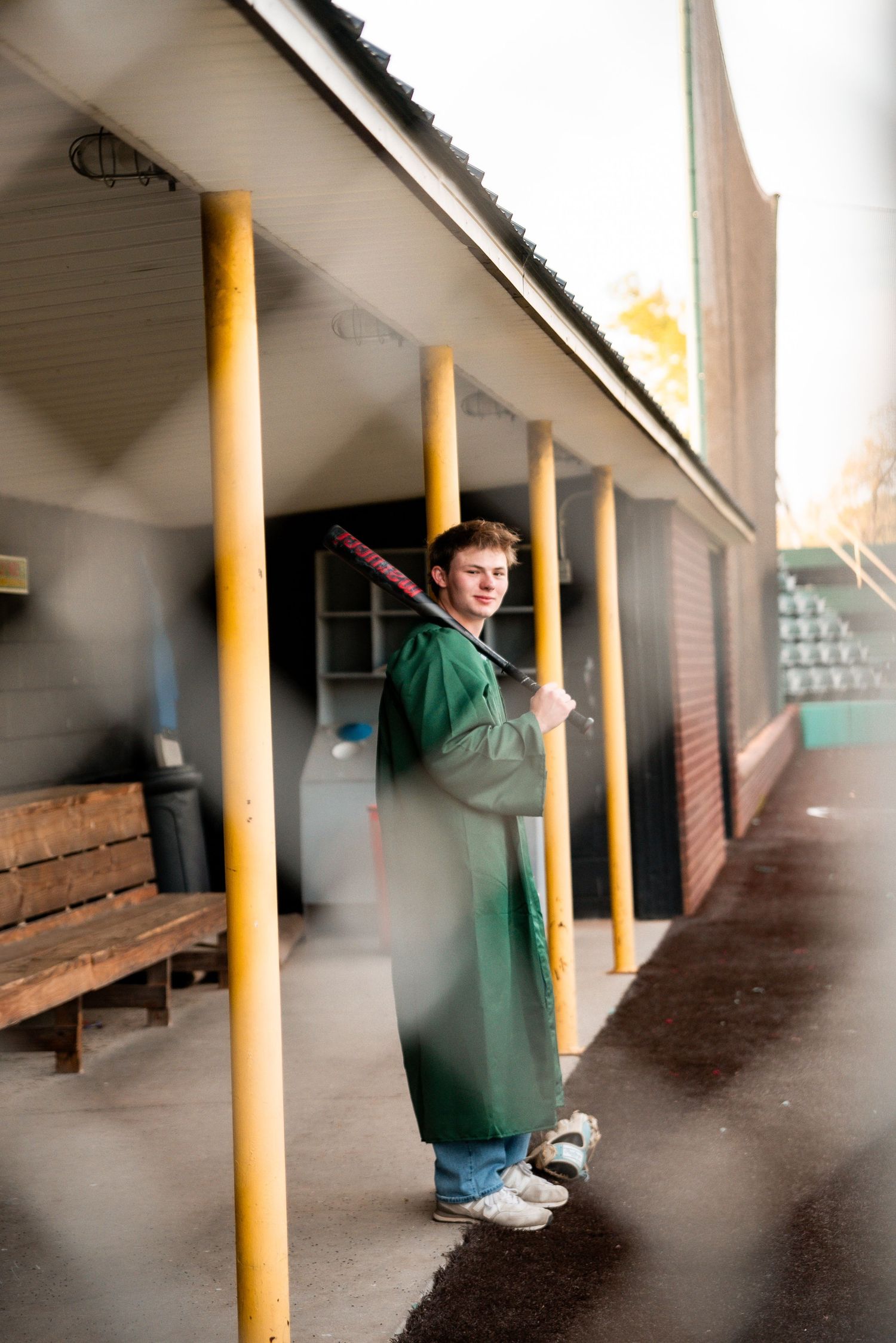 Young man in green raincoat holding a baseball bat stands in a dugout at a baseball field.