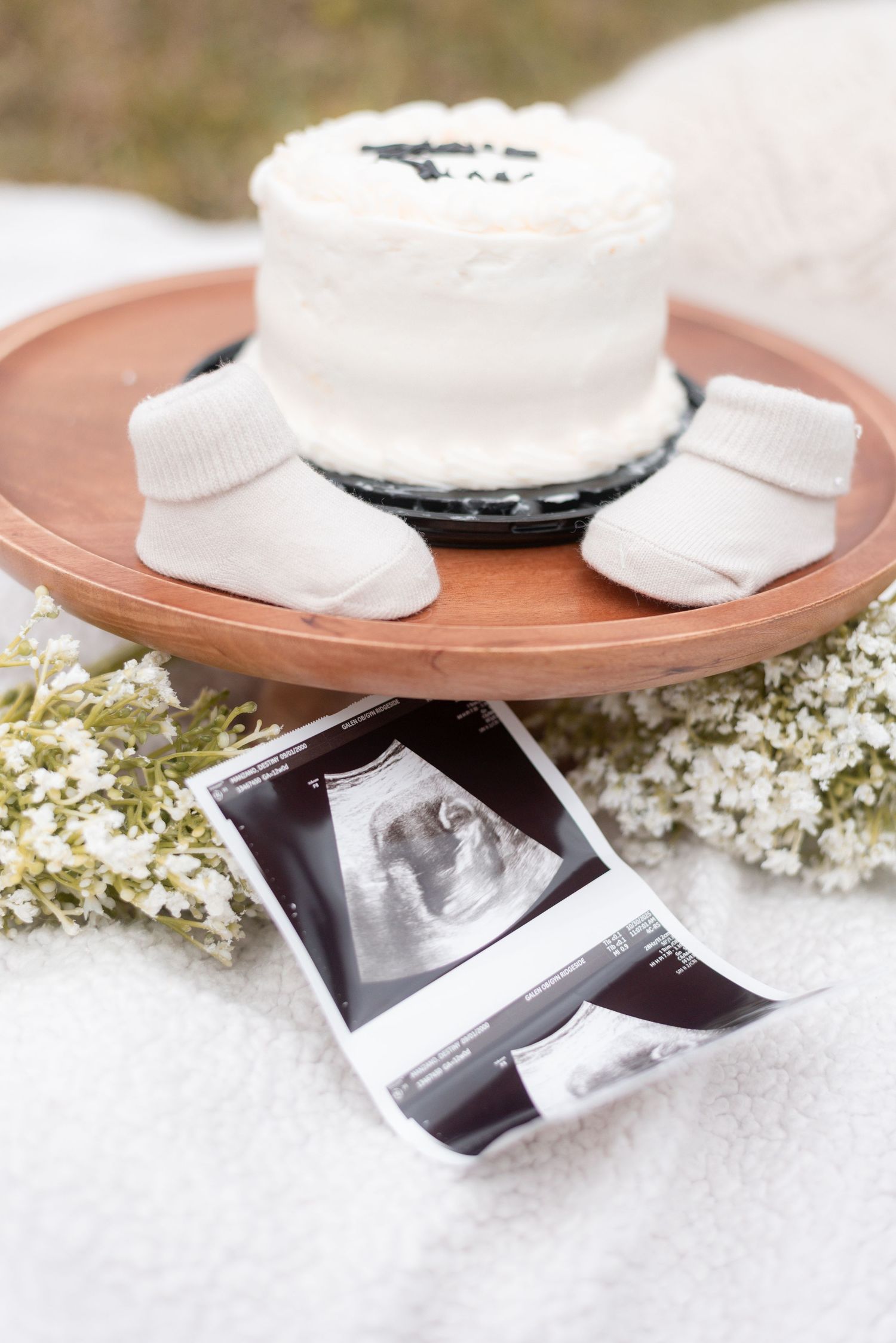 Baby booties and ultrasound photos arranged on a wooden plate with delicate white flowers.