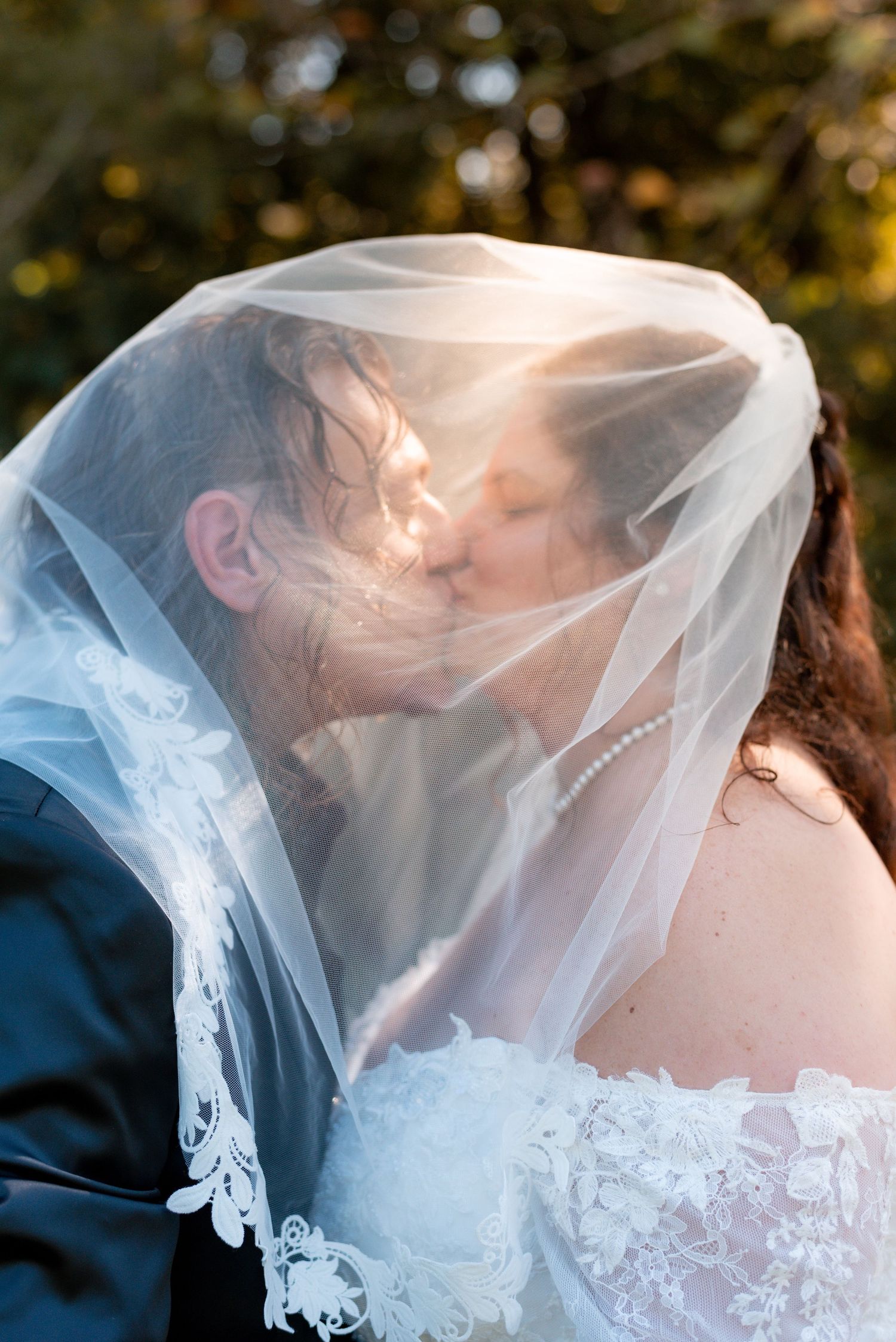 A romantic wedding kiss captured through a delicate white veil with soft bokeh lighting in the background.