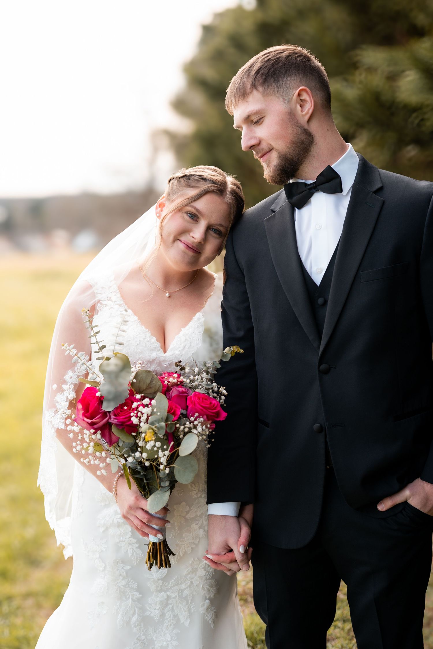 A romantic outdoor wedding portrait with a couple holding a red and white bouquet.