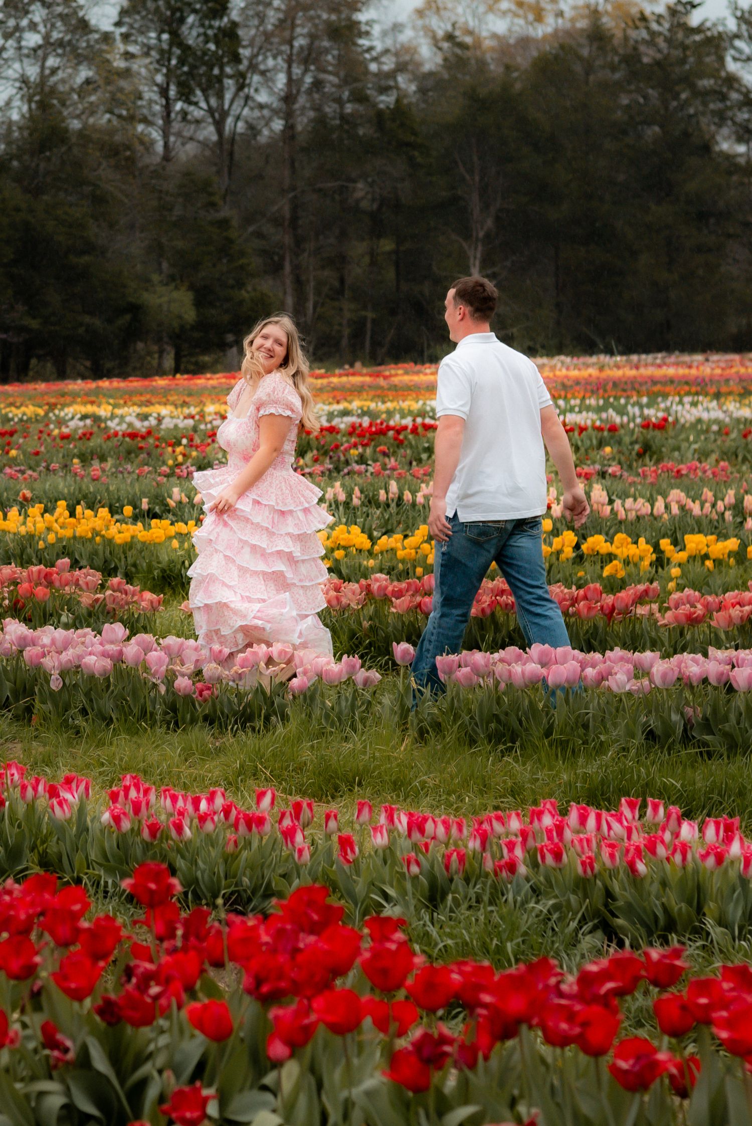Couple walking through vibrant tulip field, woman in pink ruffled dress smiling back at man in white shirt and jeans.
