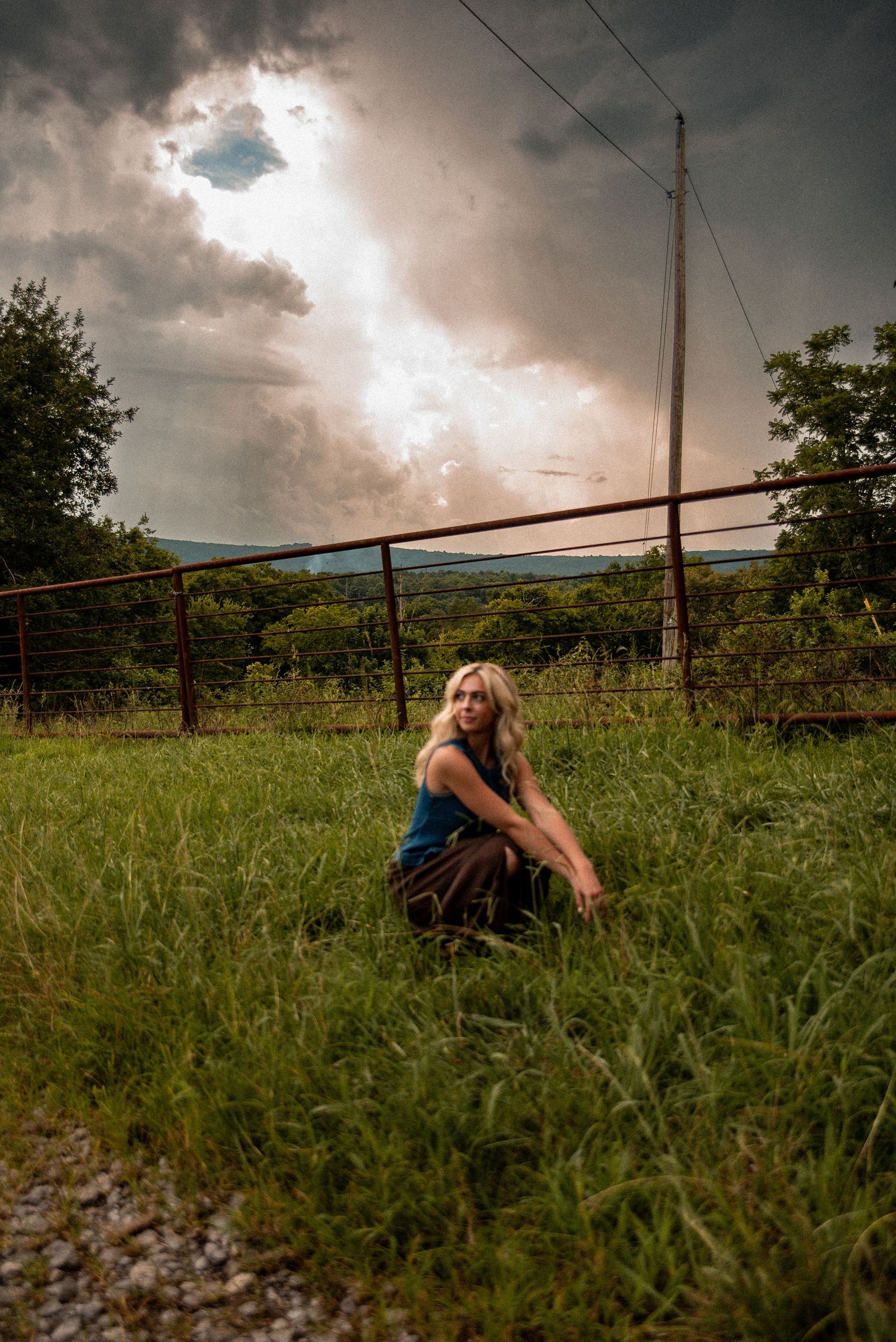 Blonde woman crouching in green grass field with dramatic stormy sky, rural fence and trees in background.
