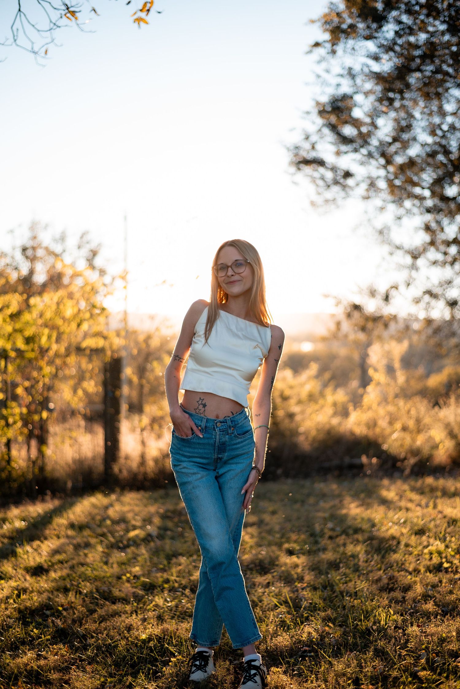 Casual outdoor portrait in jeans and white top during golden hour with trees in background.