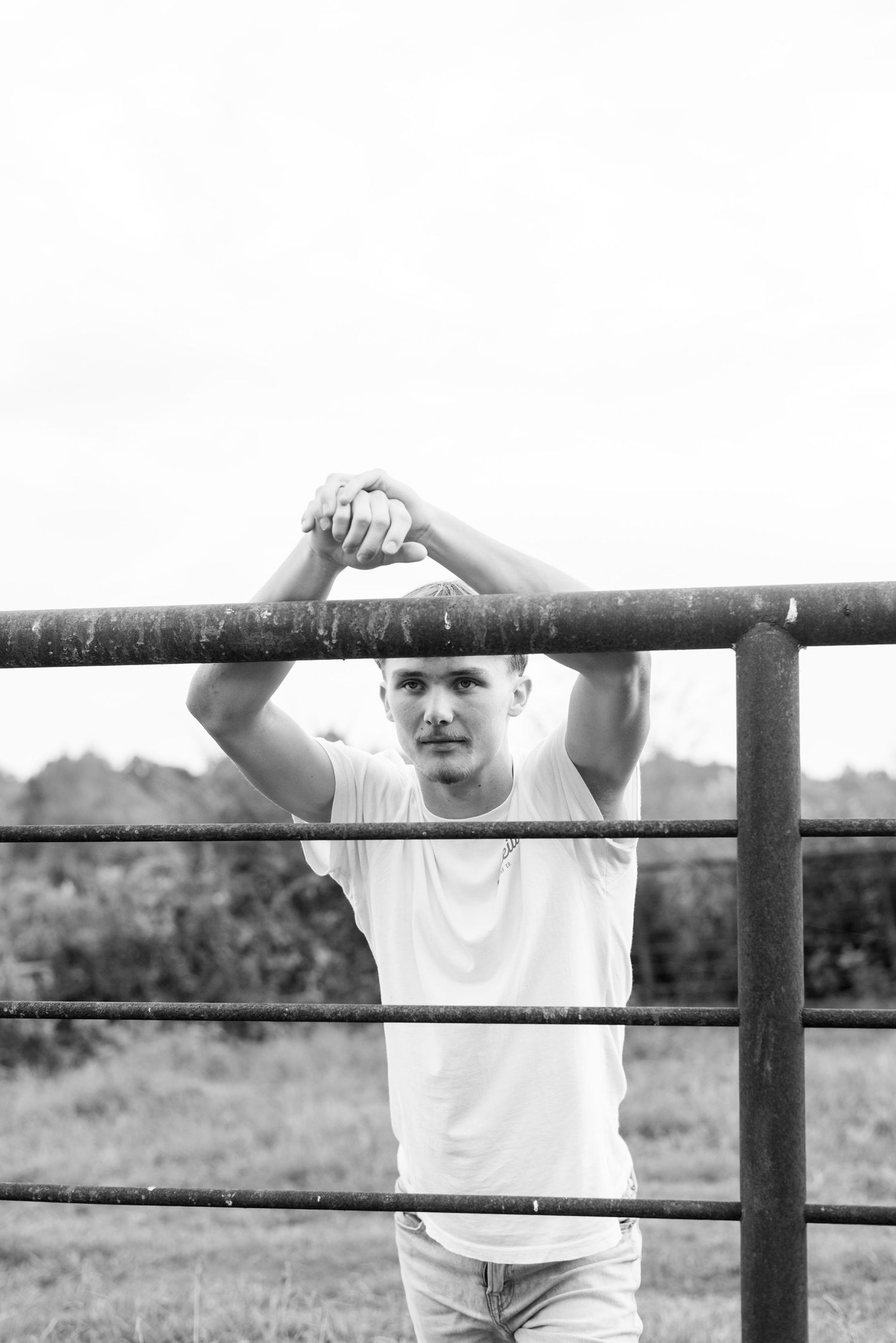 Person in white t-shirt leans on metal fence railing in rural setting.