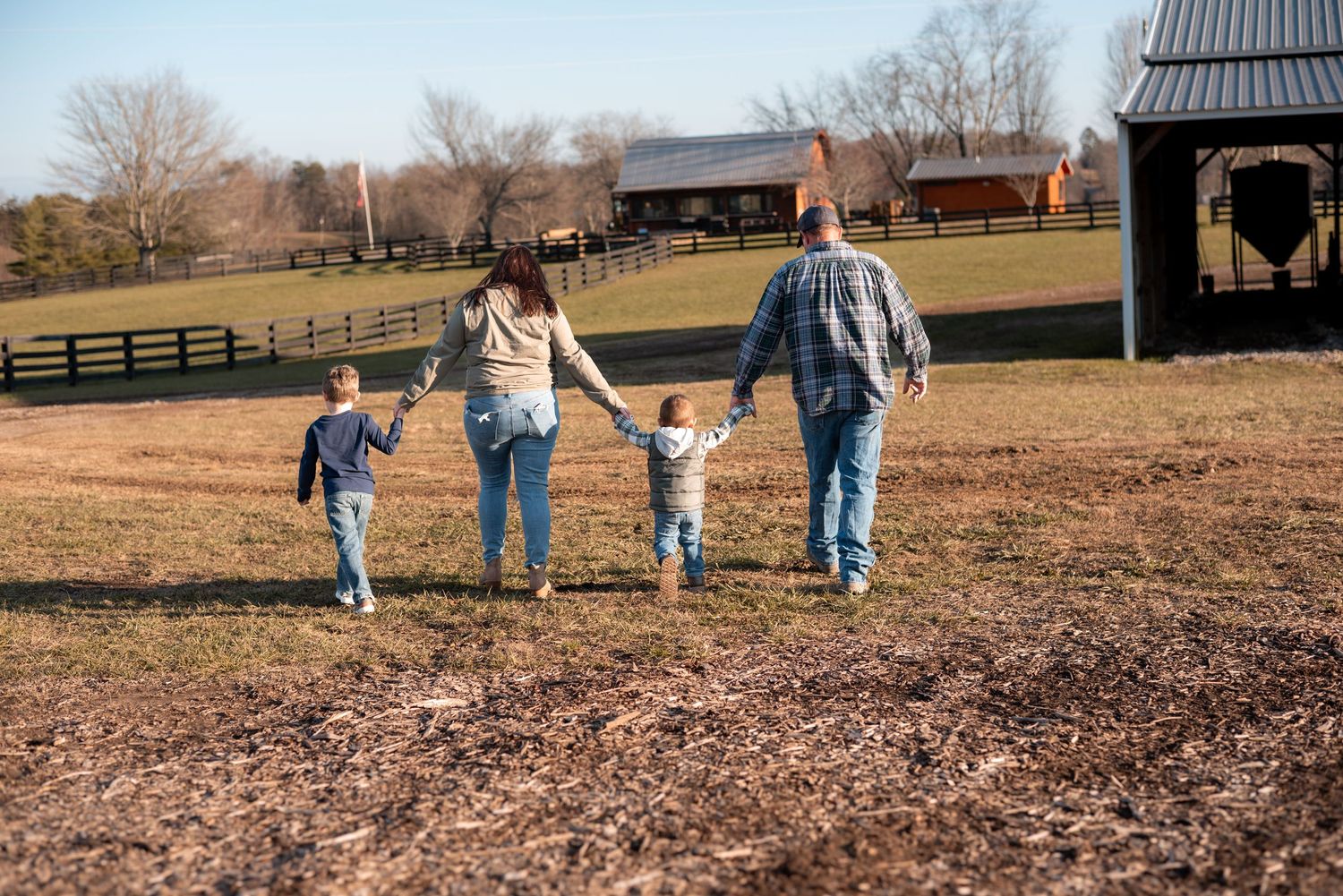 Family walking together across an open field with barns and fences in the background.