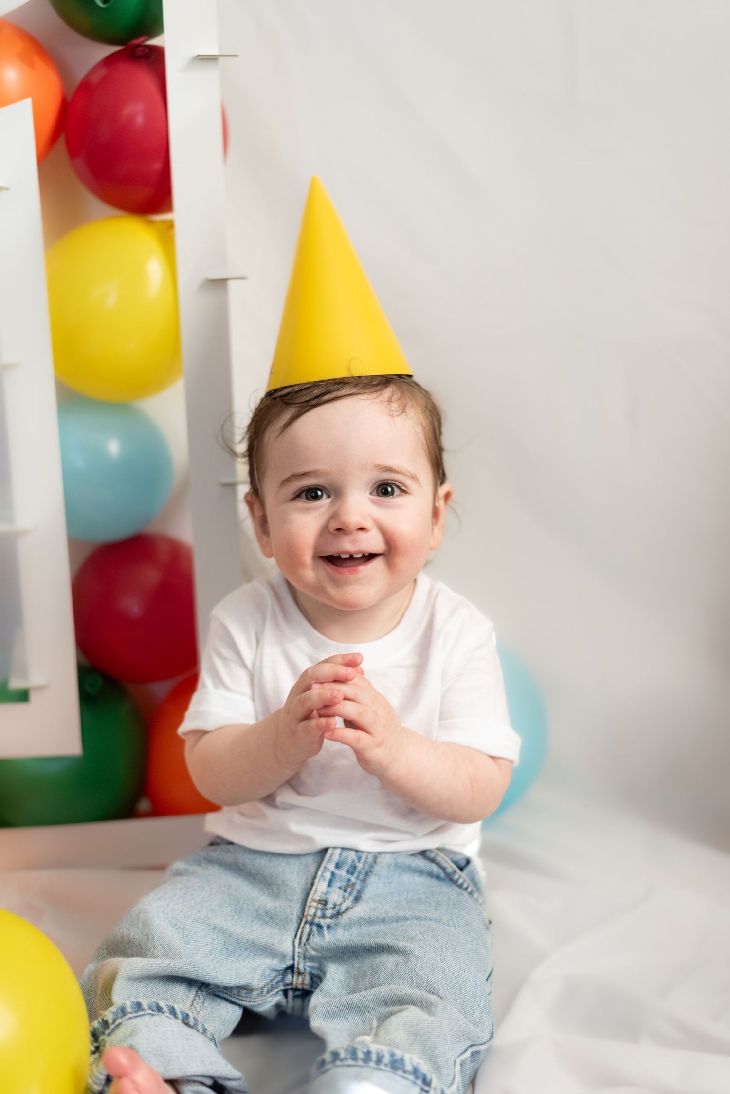 A smiling toddler in a yellow party hat sits among colorful balloons.