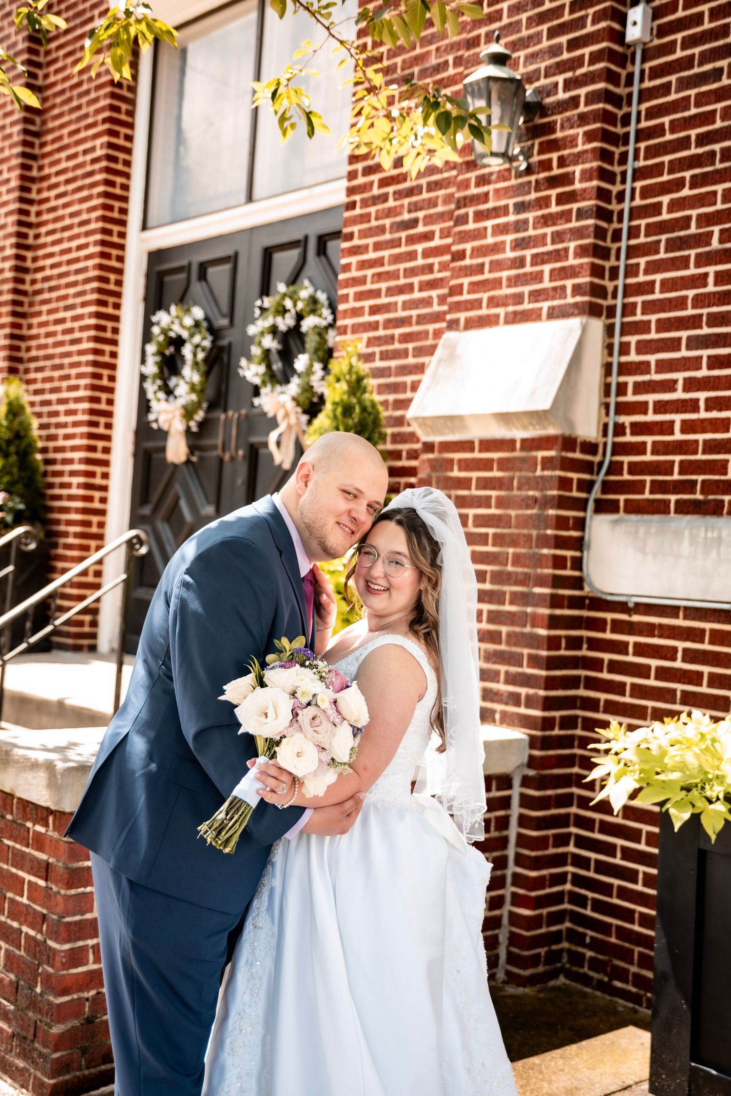 Bride and groom share a romantic moment outside a brick church, bride holding a pink and white bouquet in a white gown.