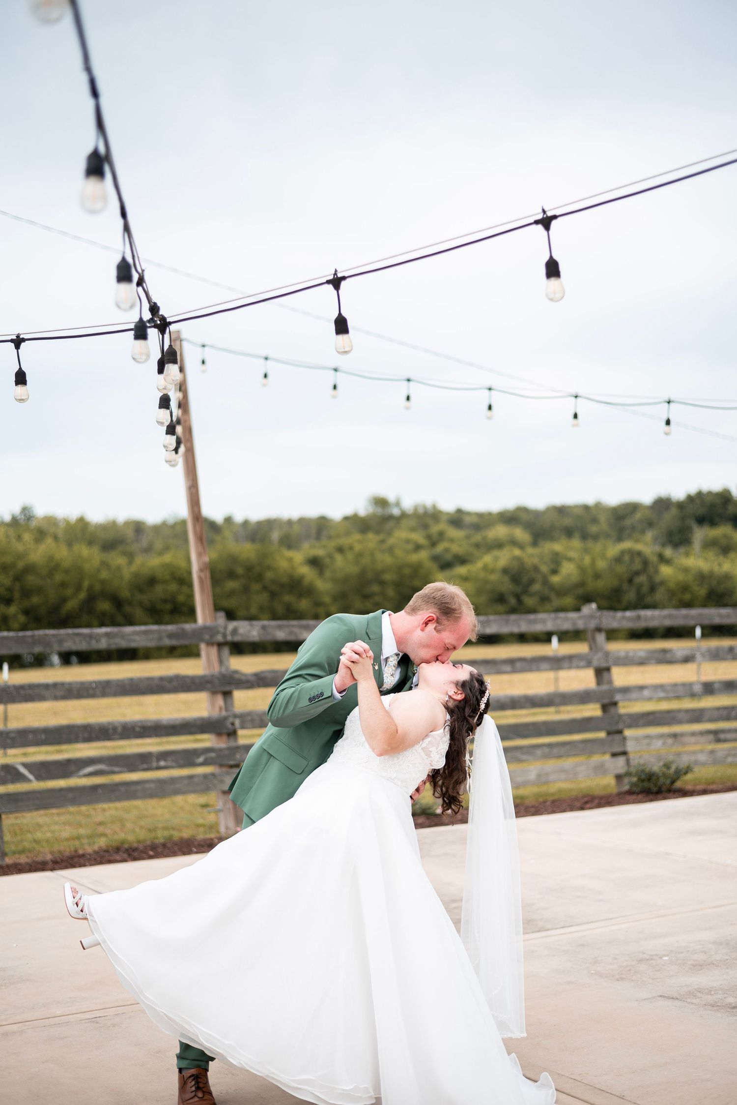 A groom in a green suit dips his bride in a white wedding dress outdoors, surrounded by string lights.