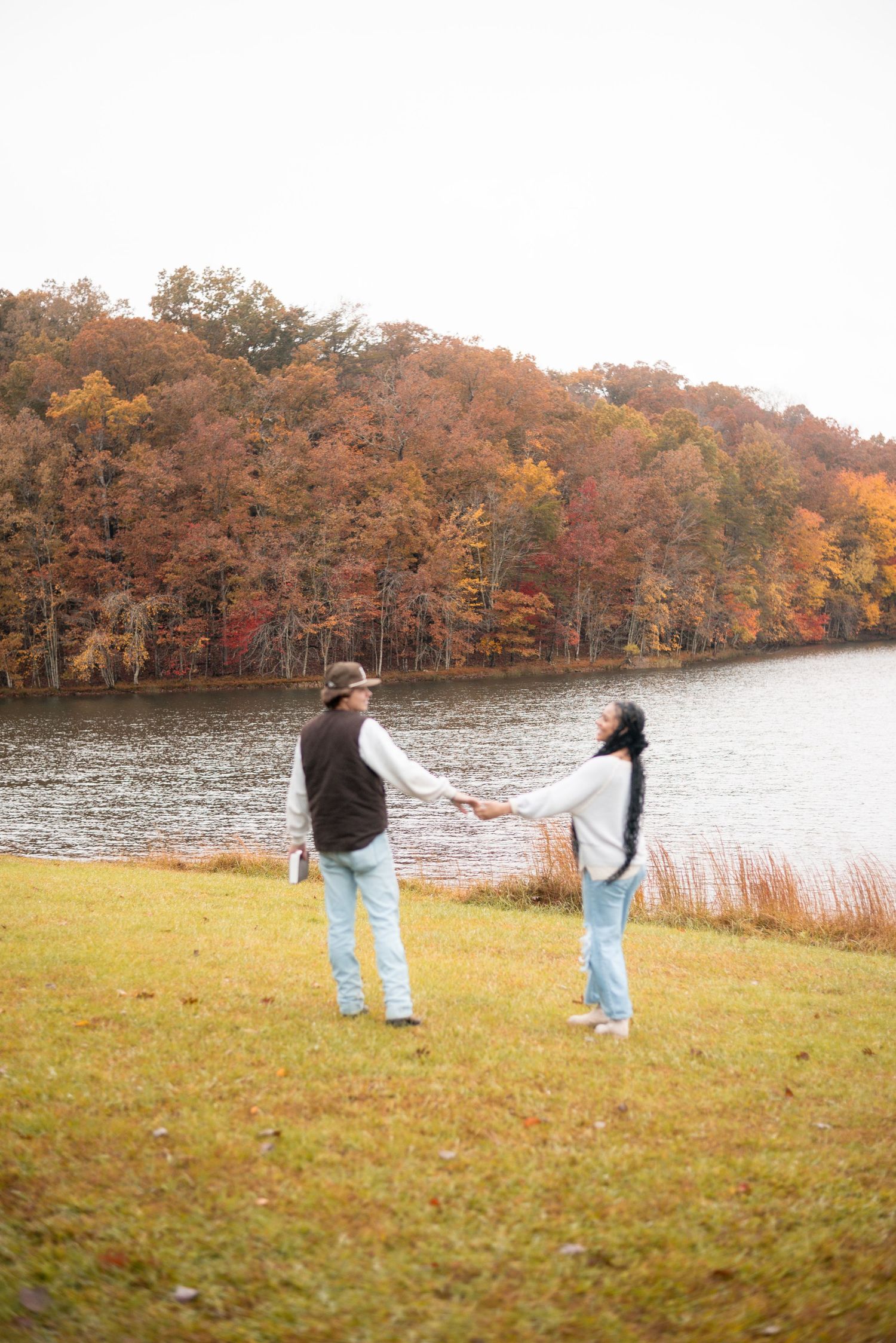 Two people playfully interact on a grassy field with fall-colored trees and a lake in the background.