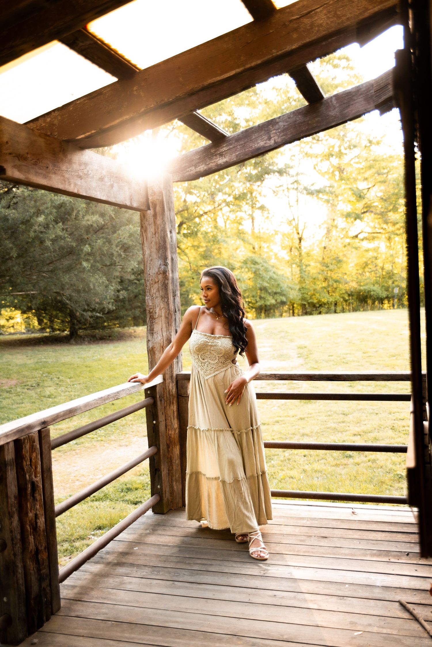 Woman in elegant cream maxi dress standing on rustic wooden porch at golden hour sunset.