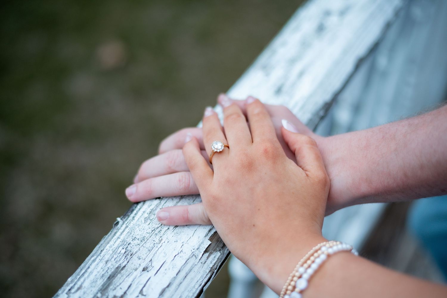 Two hands resting on a weathered wooden fence, one adorned with a diamond engagement ring and pearl bracelet.