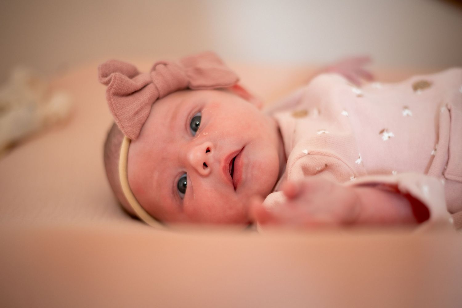 Newborn baby wearing pink outfit with bow headband looking at camera.