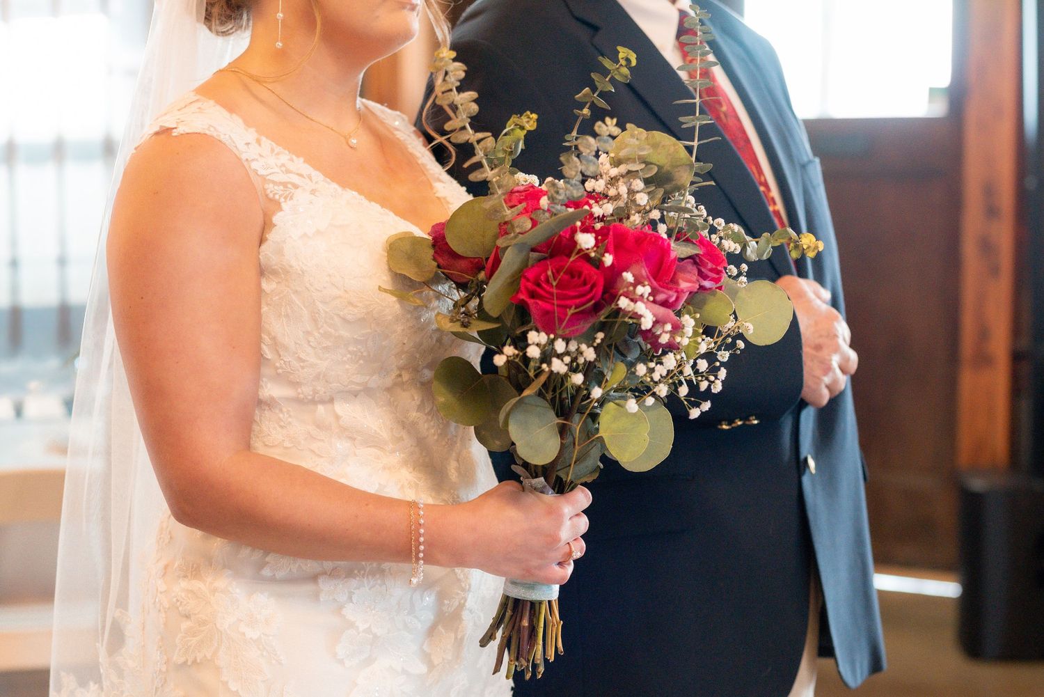 A close-up of a bridal bouquet featuring red roses and greenery with white lace dress detail.