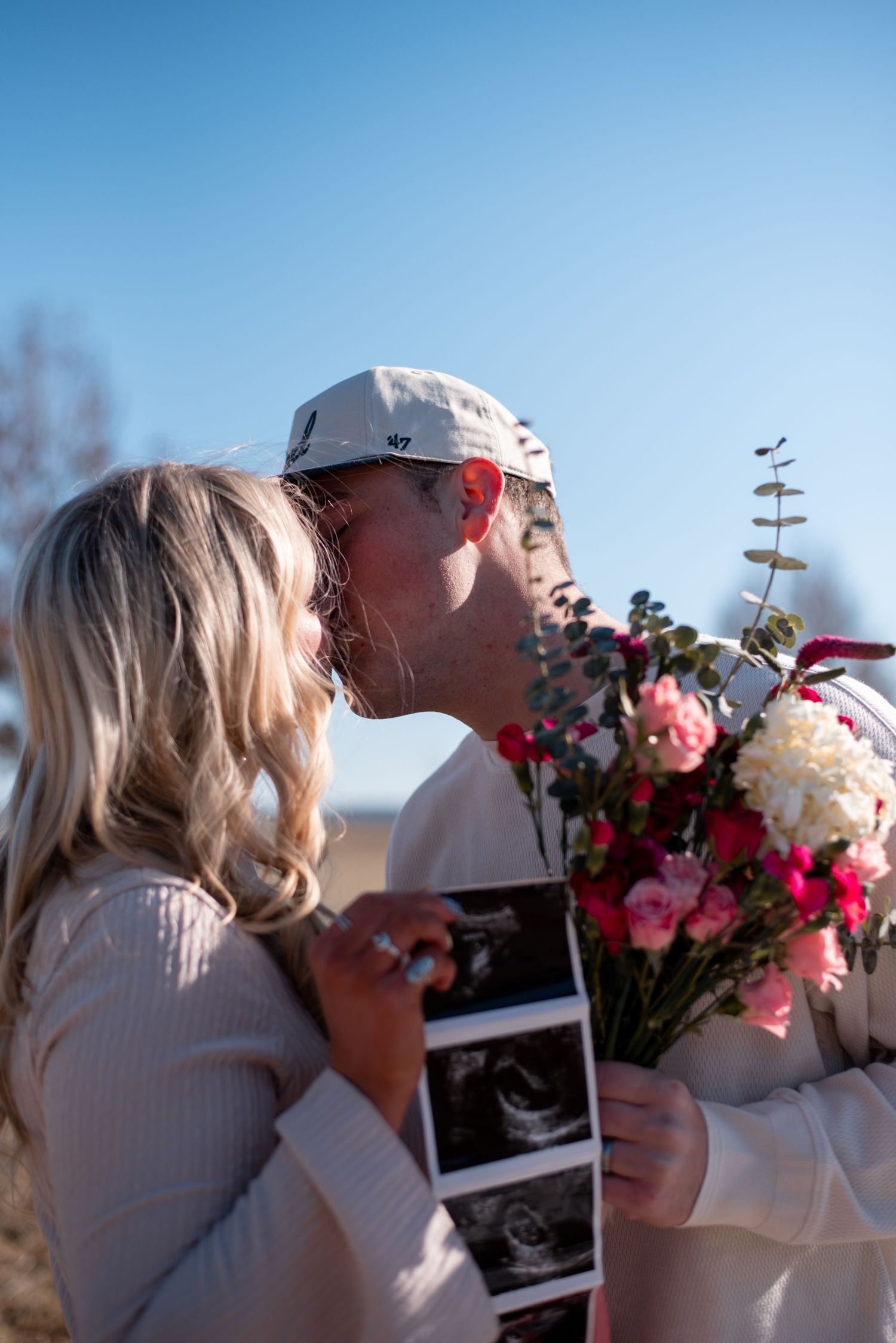 A couple shares a romantic moment outdoors while holding flowers and an ultrasound photo.