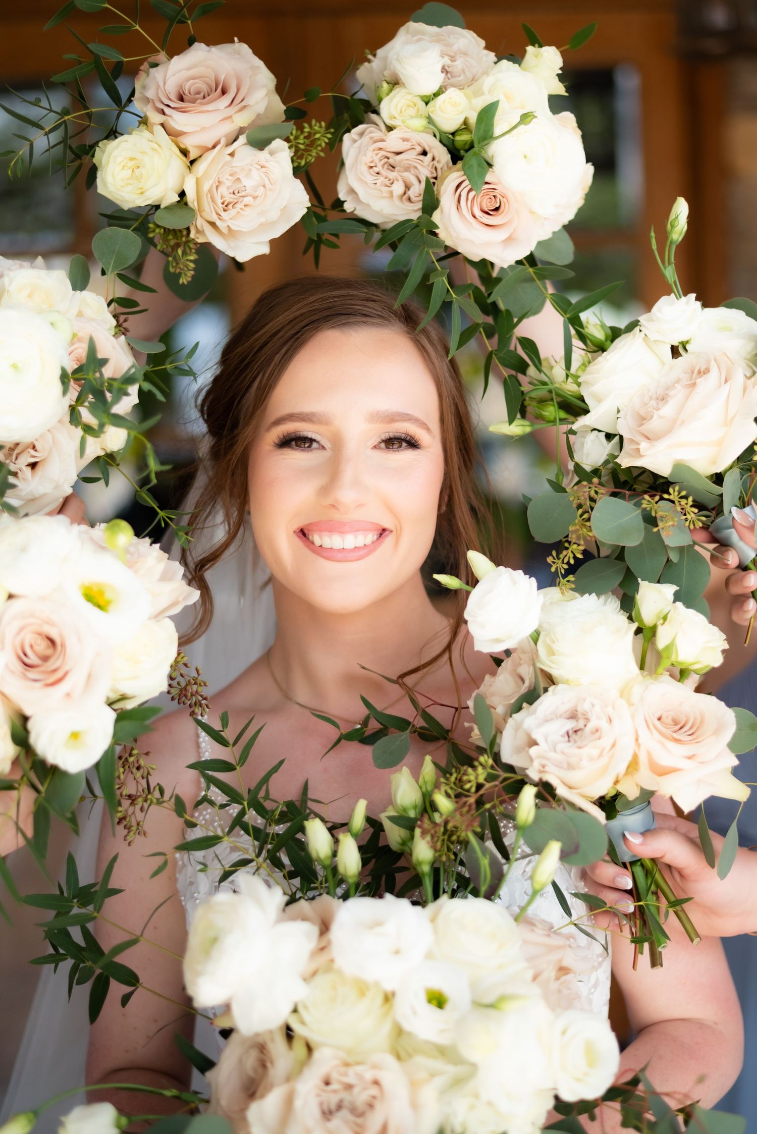 Smiling bride surrounded by lush blush and white rose bouquets with eucalyptus greenery framing her face on her wedding day.