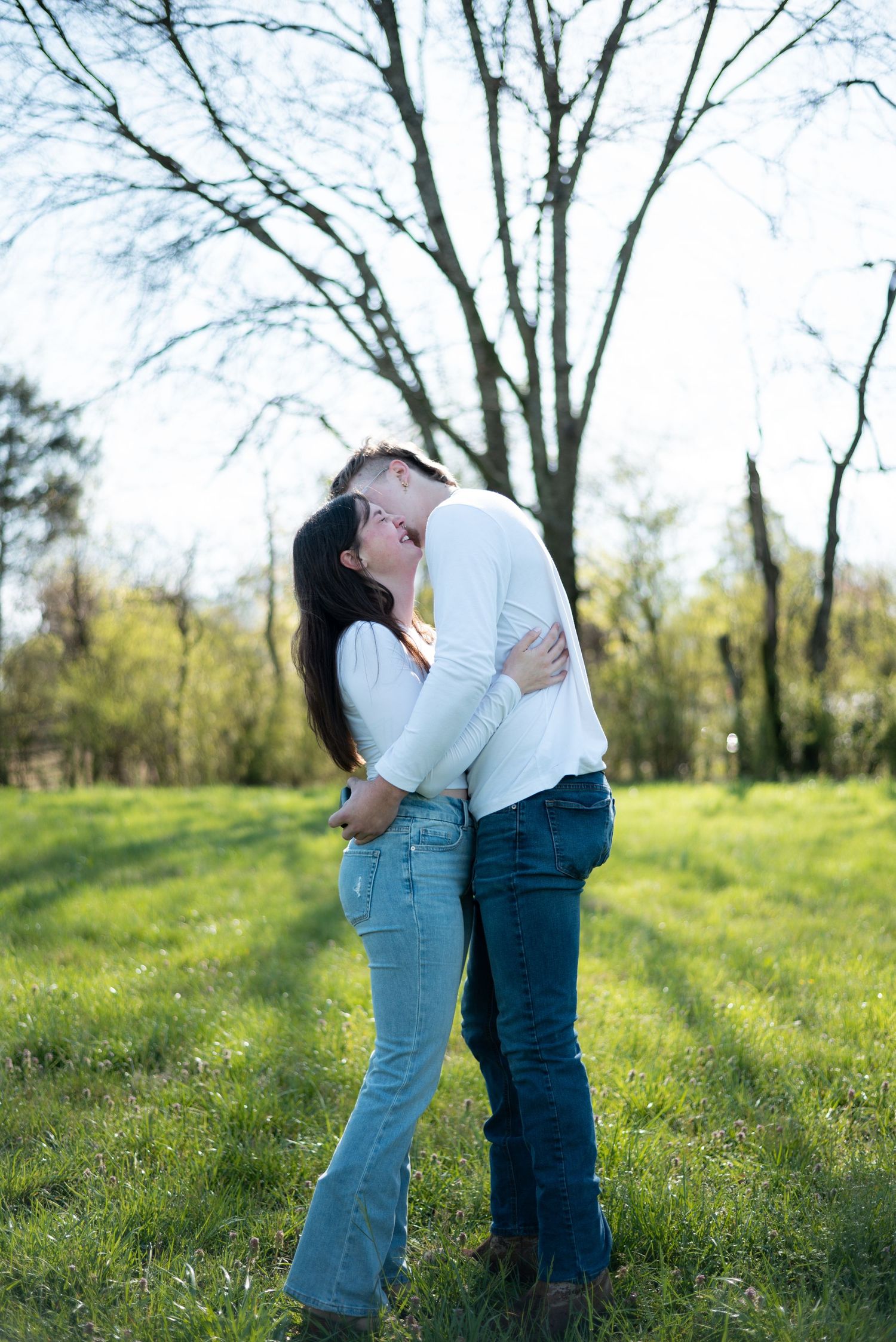 Couple sharing a romantic kiss outdoors in a green park, both wearing white tops and blue jeans on a sunny day.