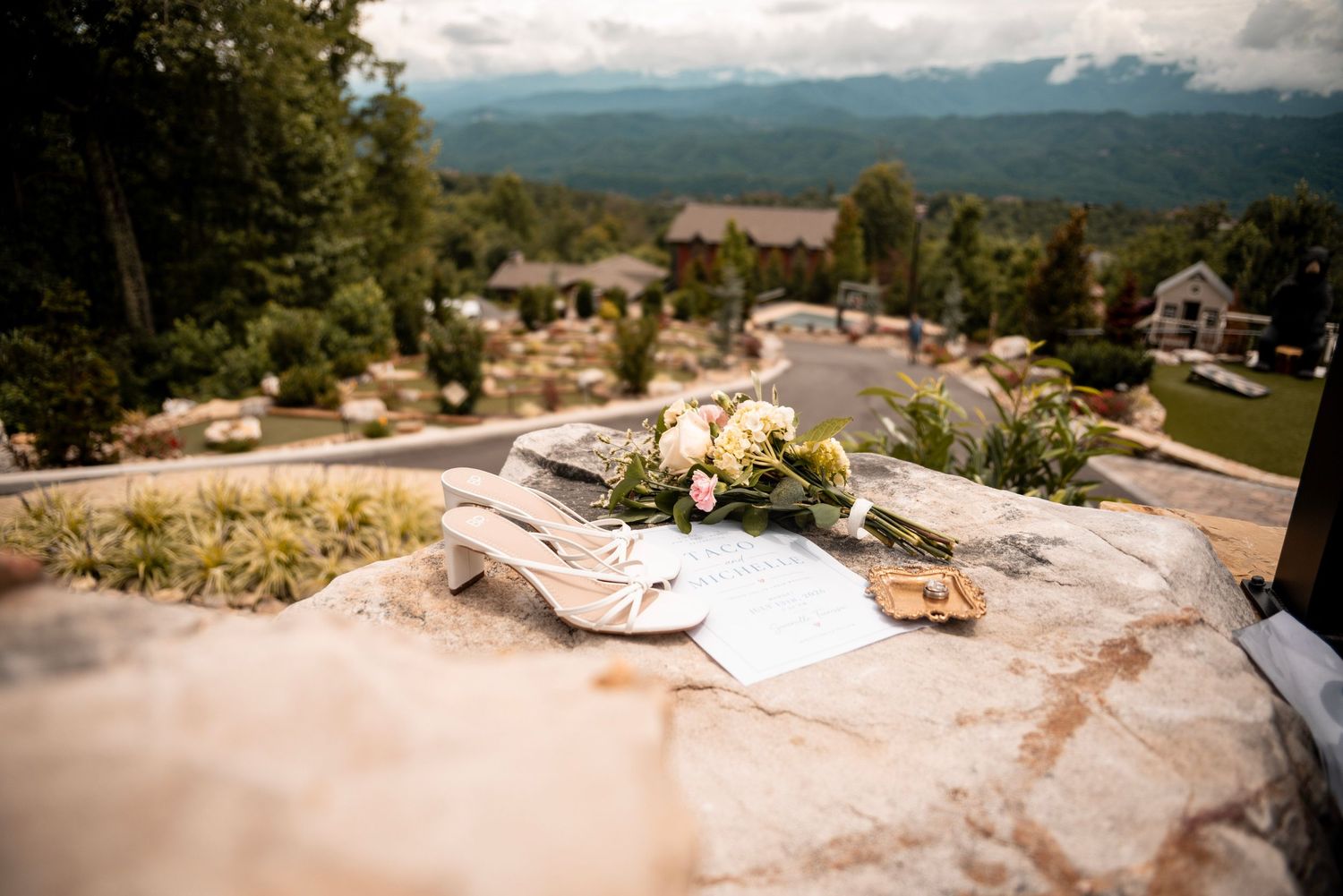 Wedding shoes, bouquet, and vows on a stone ledge with mountain views and venue in background.
