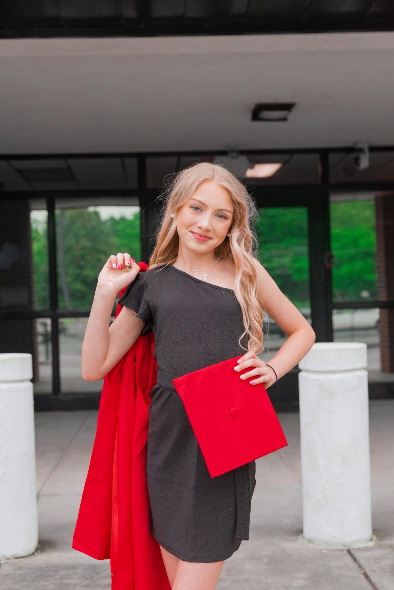 Graduate in black dress holding red graduation gown outside building.