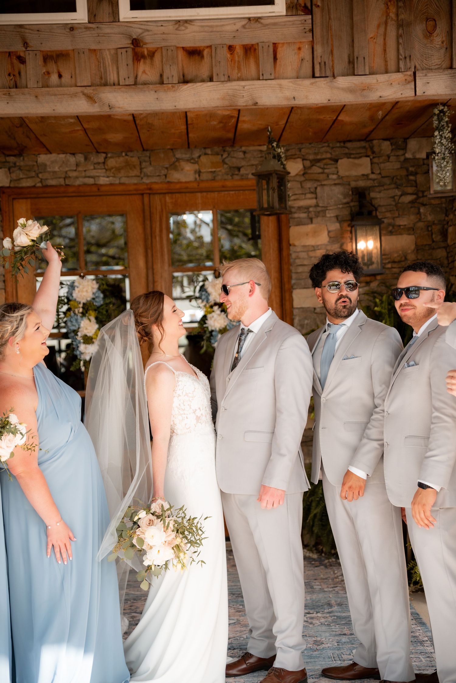 Wedding party in gray suits and blue bridesmaid dress pose outside rustic stone venue with bride holding white bouquet.