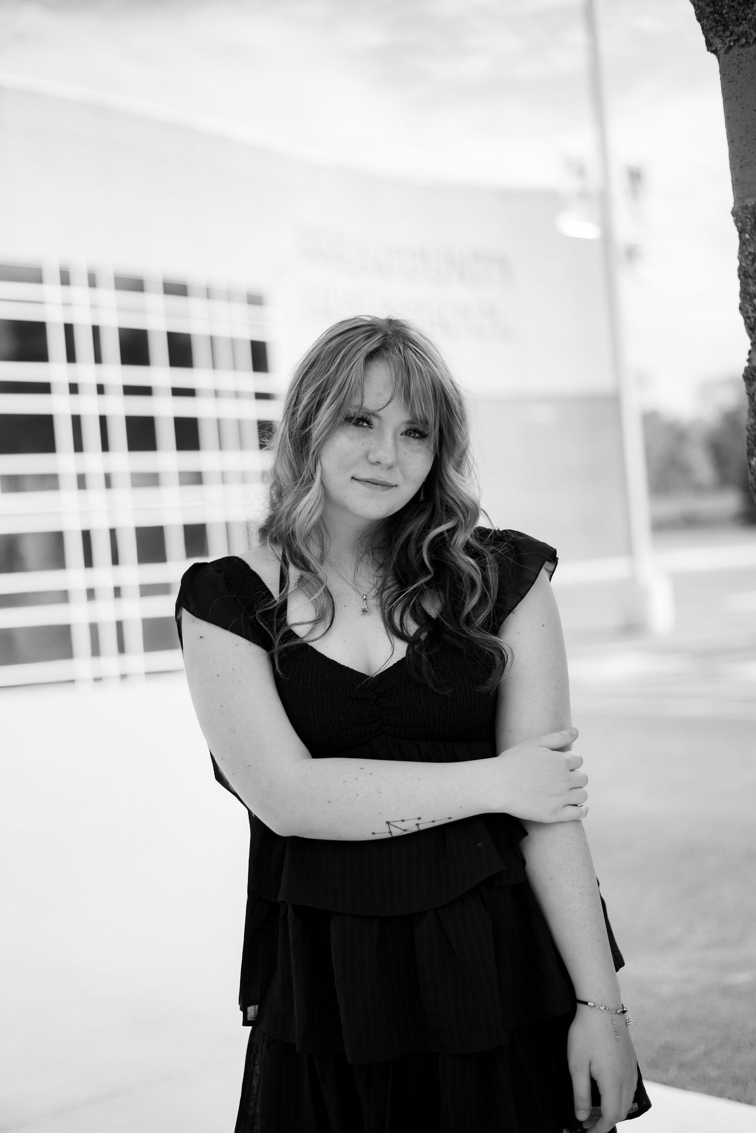 Young woman in black dress posing outdoors with arms crossed in black and white photo.