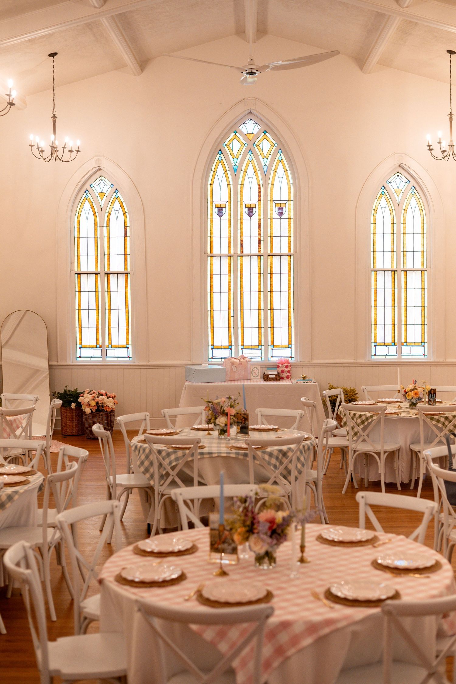 Elegant chapel venue with stained glass windows, white chairs, and pink gingham tablecloths set for a wedding reception.