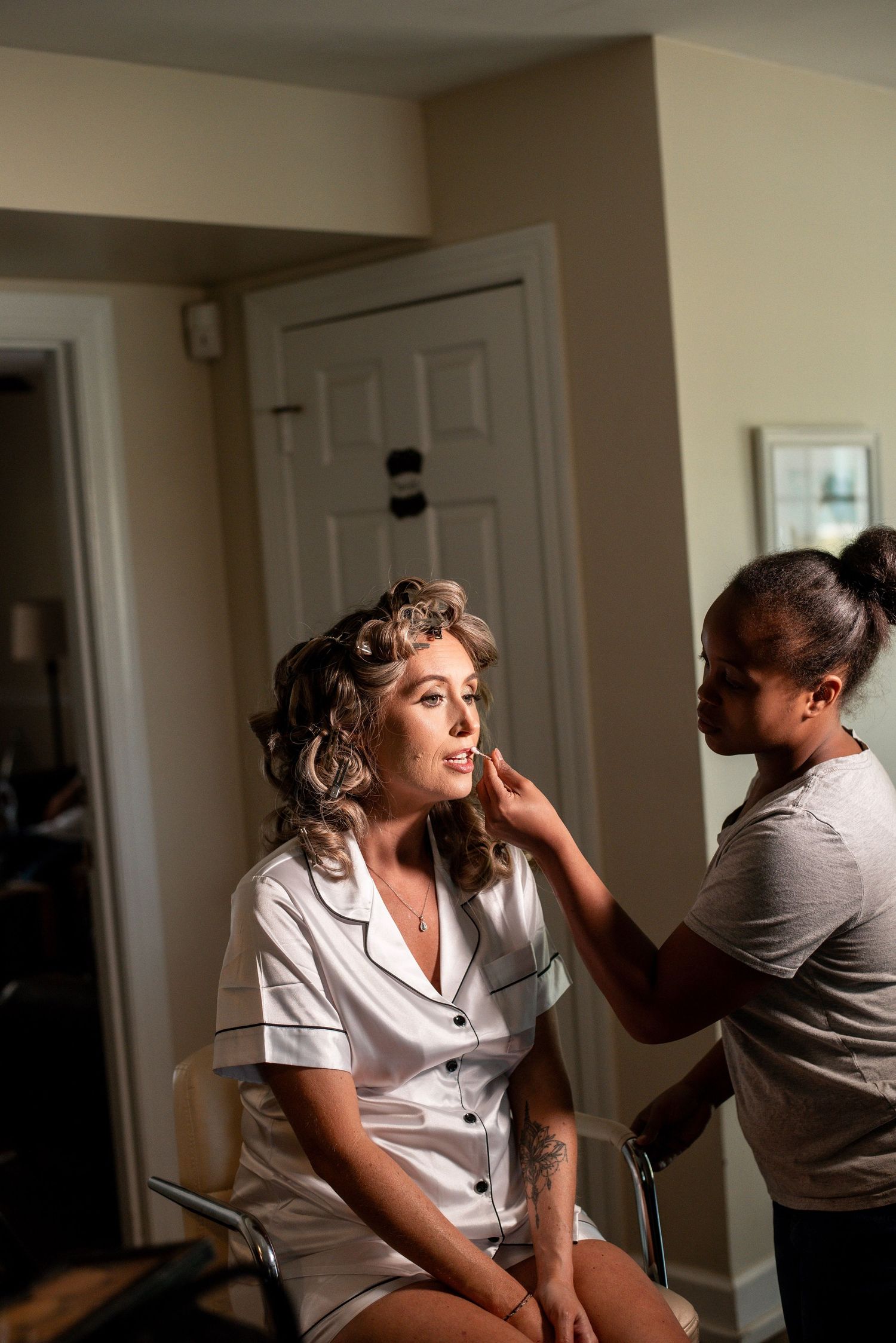 Bride getting makeup applied by artist while sitting in chair with hair in curlers before wedding.