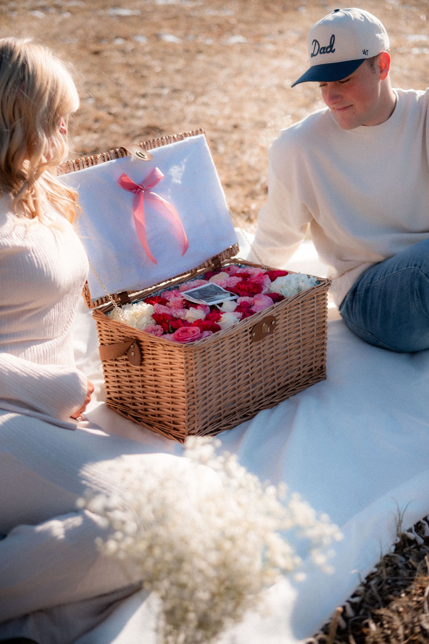 Wicker basket filled with pink flowers and gifts on white blanket in sunlight.