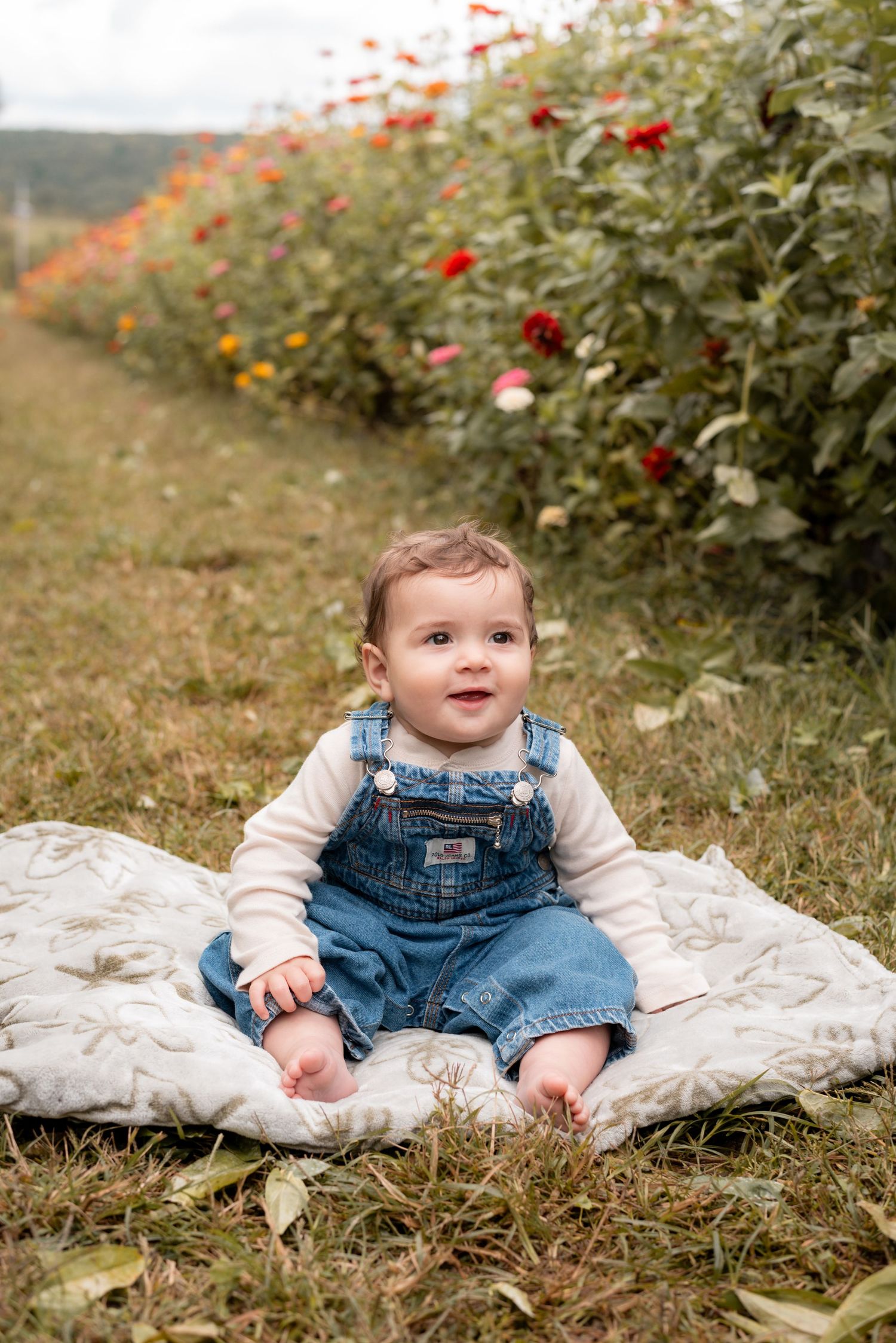 Baby in denim overalls sits on white blanket surrounded by colorful flowers in garden.