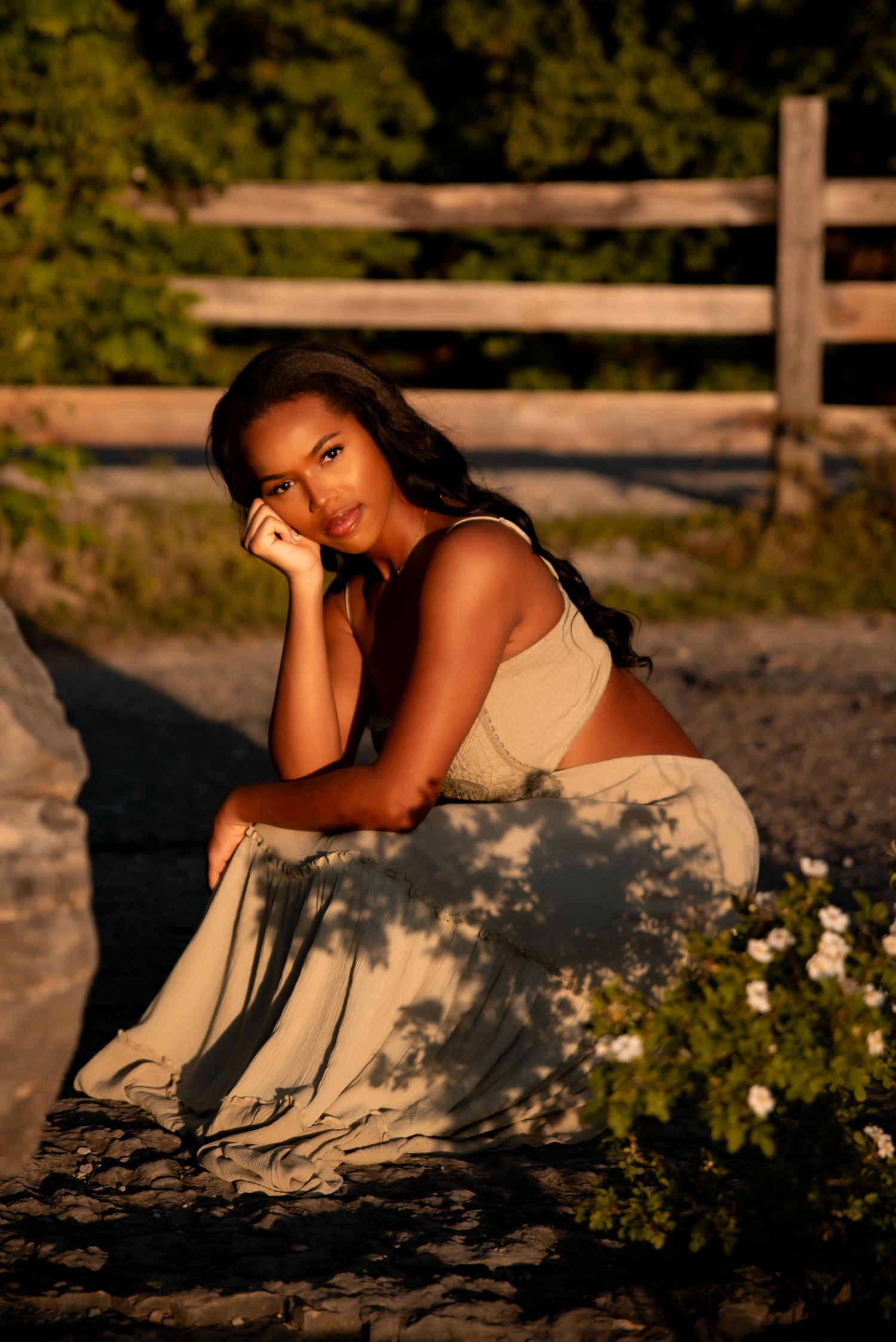 Young woman in crop top and maxi skirt poses on rocks at golden hour near rustic wooden fence.