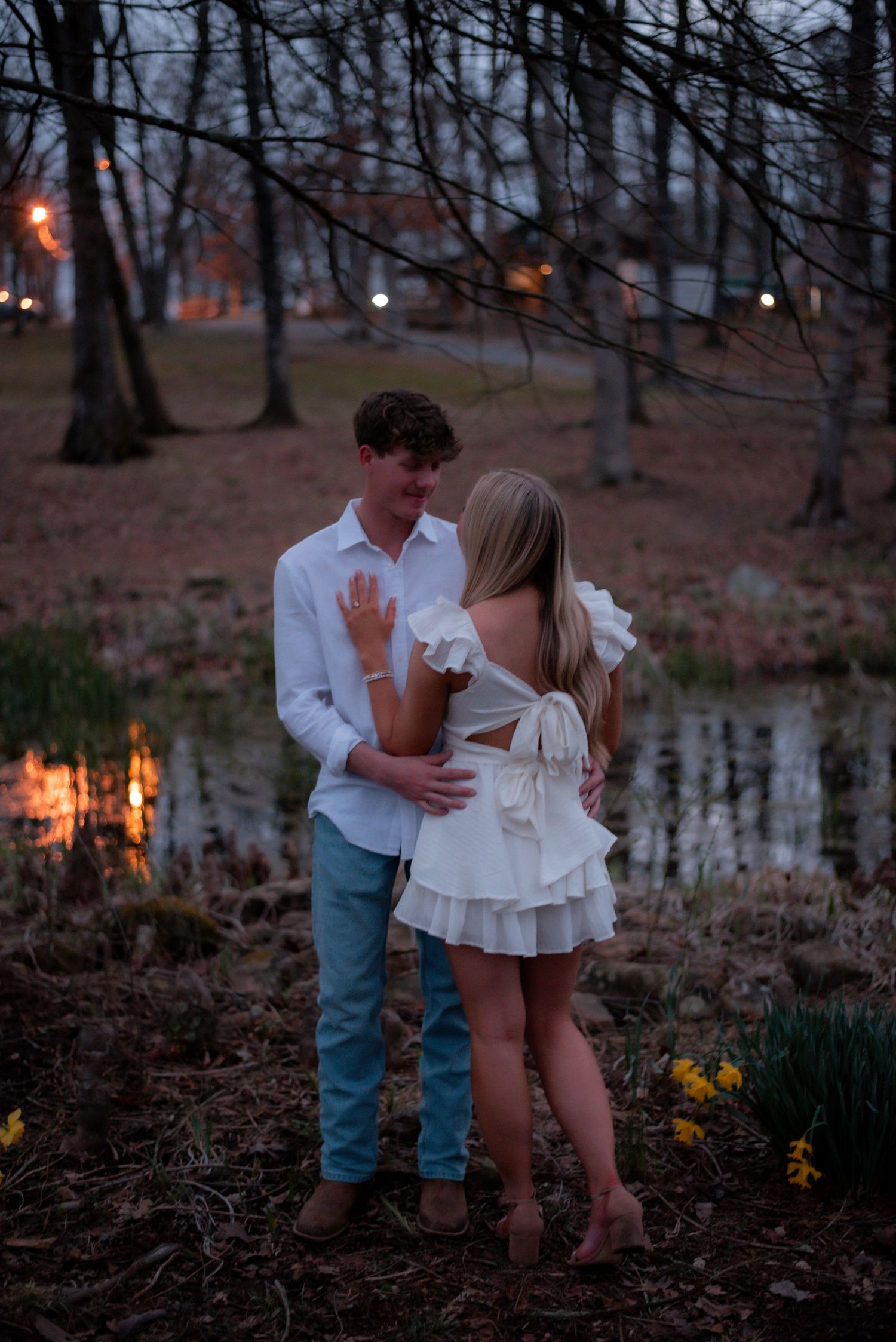 Couple in white outfits sharing a romantic moment near a pond at dusk, surrounded by bare trees.