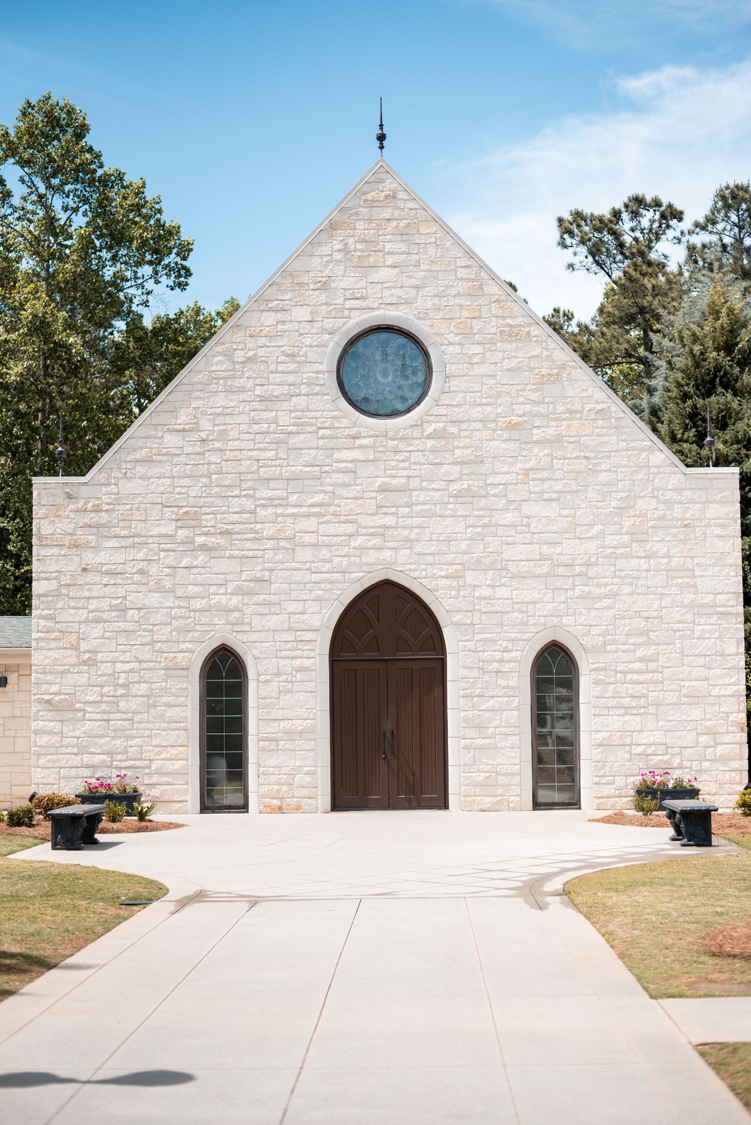 White stone chapel with gothic arched doors, round window, and tree-lined backdrop on a sunny day.