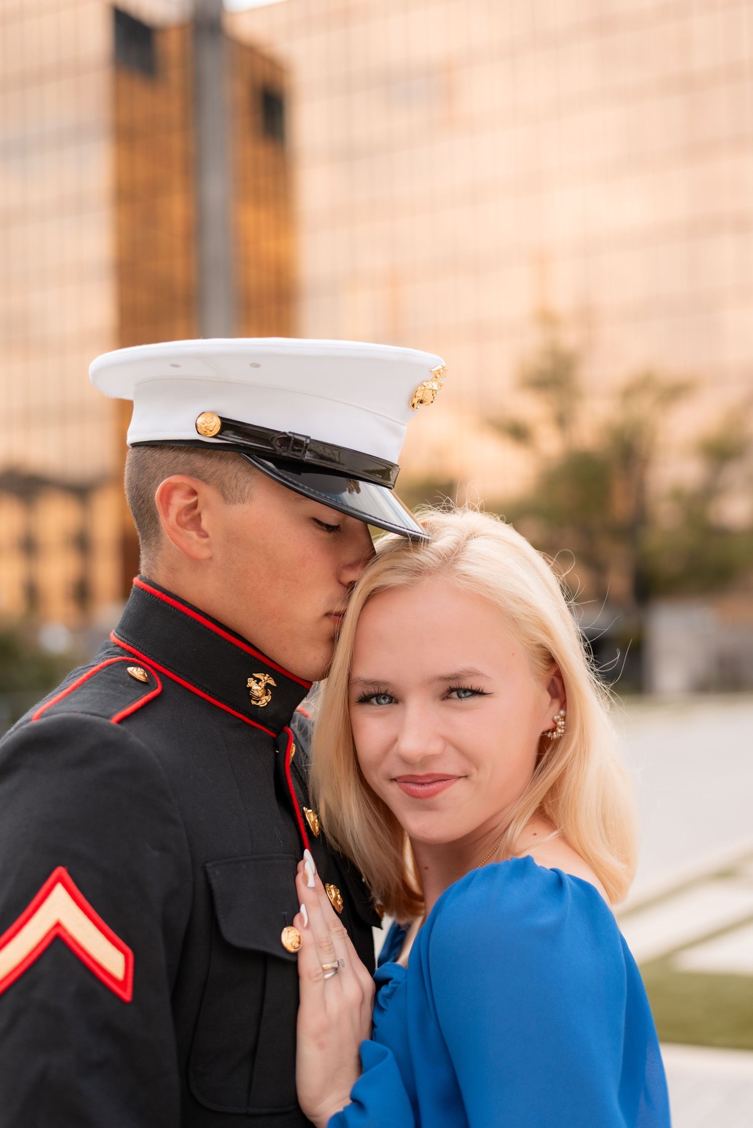 A Marine in dress uniform shares an intimate moment with someone in a bright blue dress outdoors.