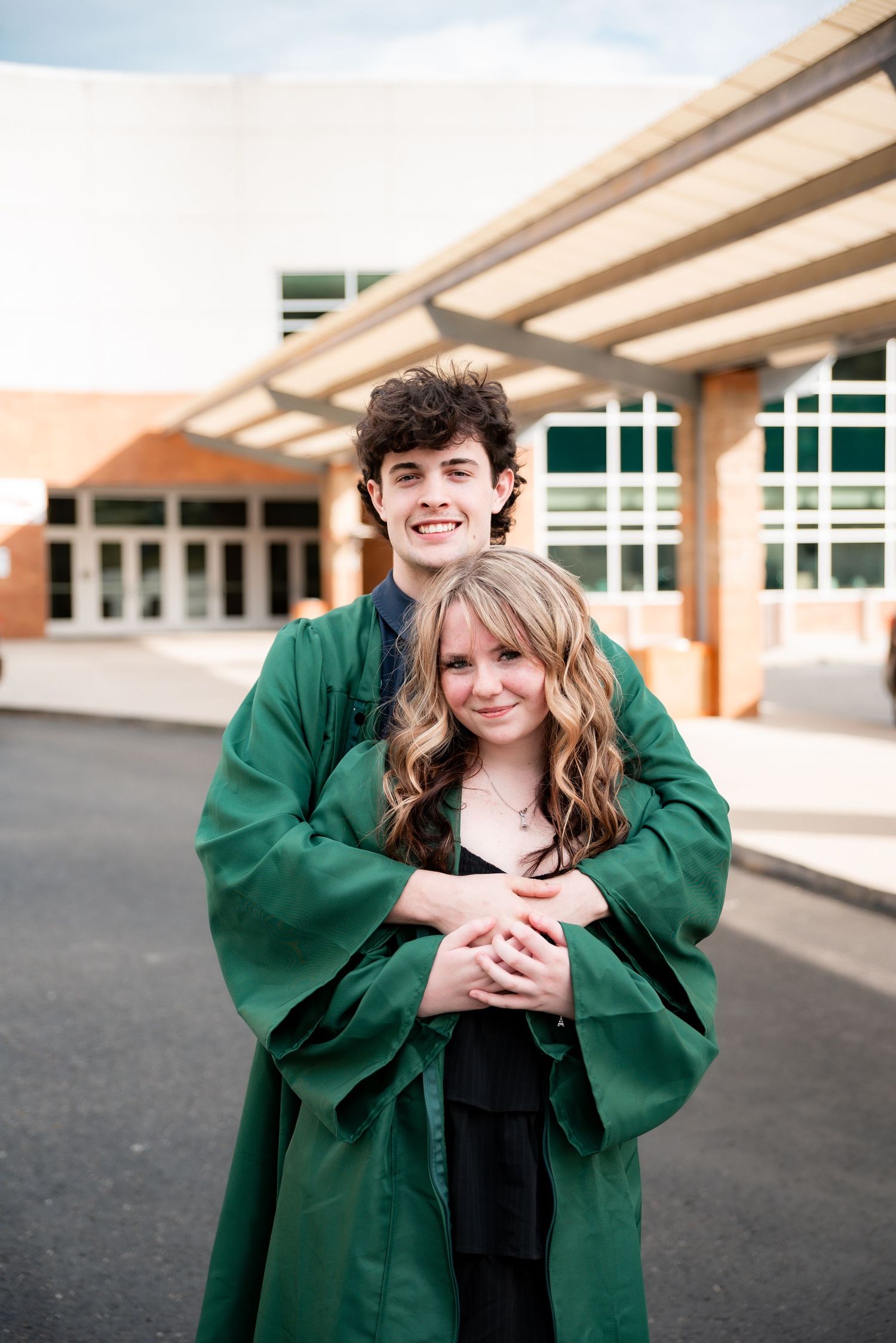 Two smiling graduates in green robes embrace outside a school building for a graduation photo.