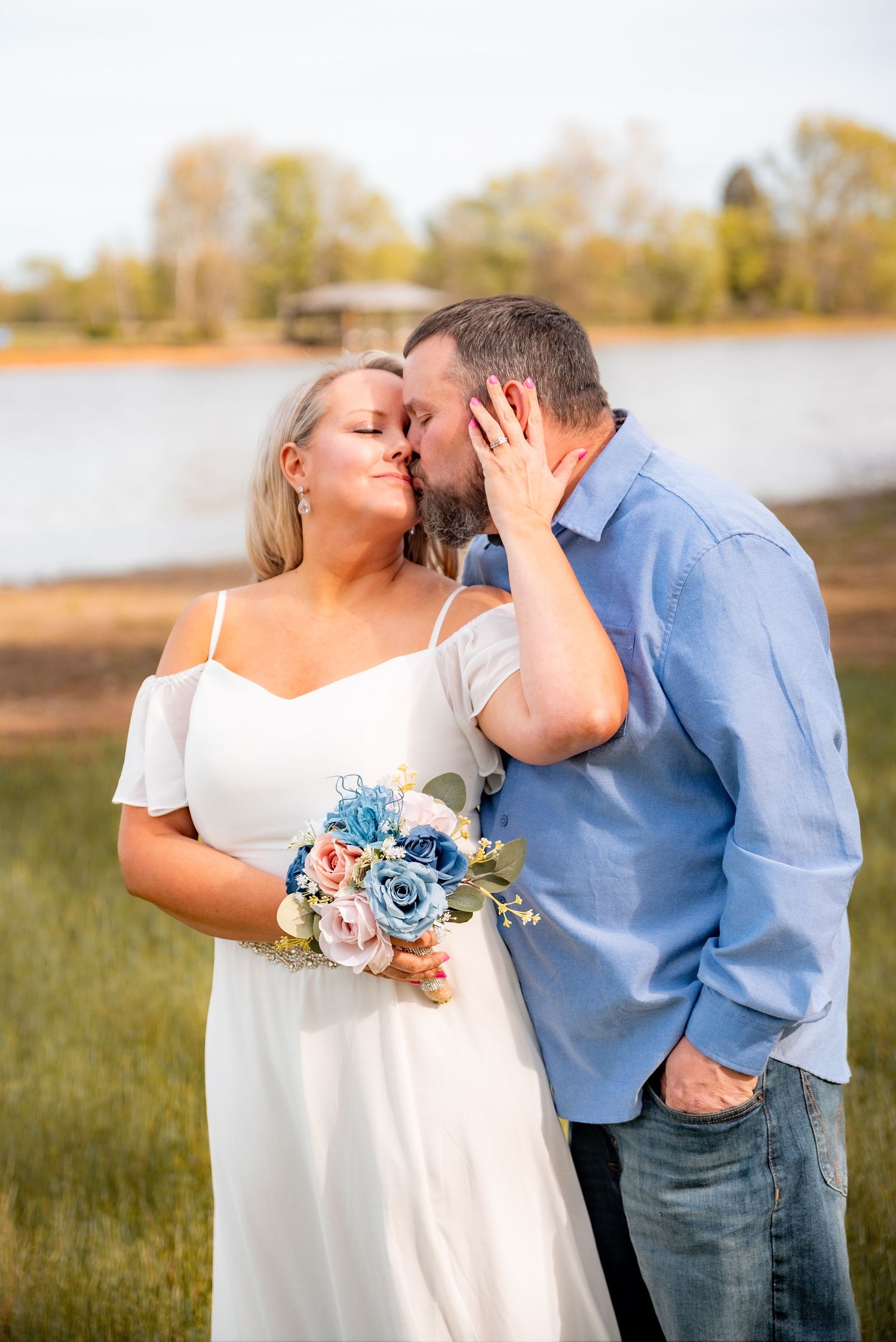 Couple sharing a romantic kiss outdoors, bride in white dress holding blue and pink bouquet near a serene lake.
