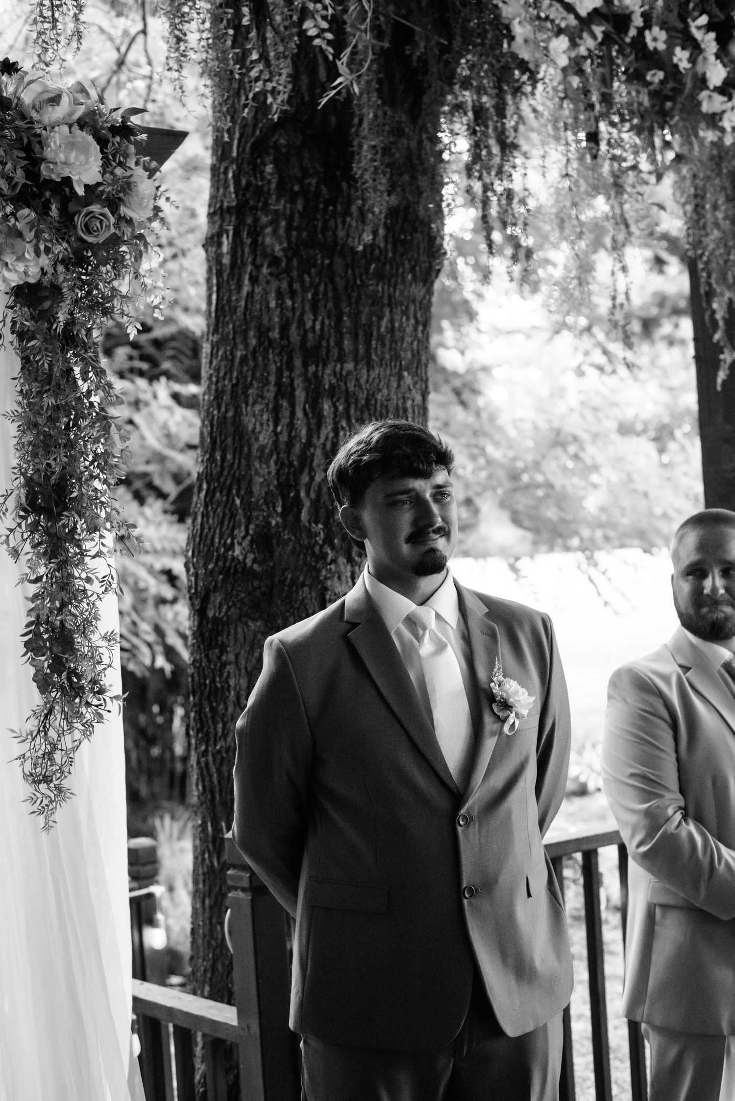 Groom in suit with boutonniere stands at wedding altar decorated with floral arrangements and greenery, black and white photo.