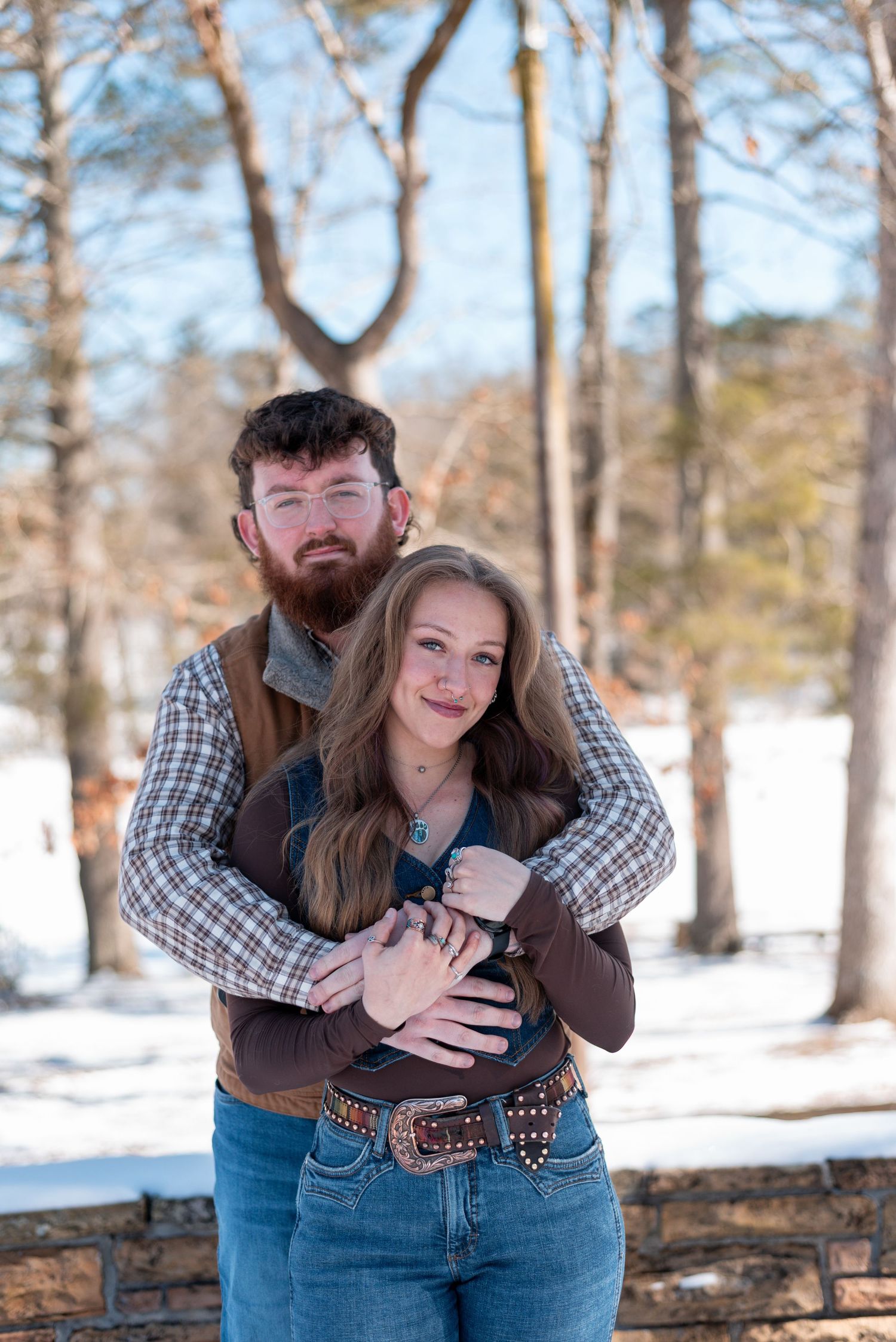 A couple in casual winter attire embracing on a snowy deck with bare trees in the background.