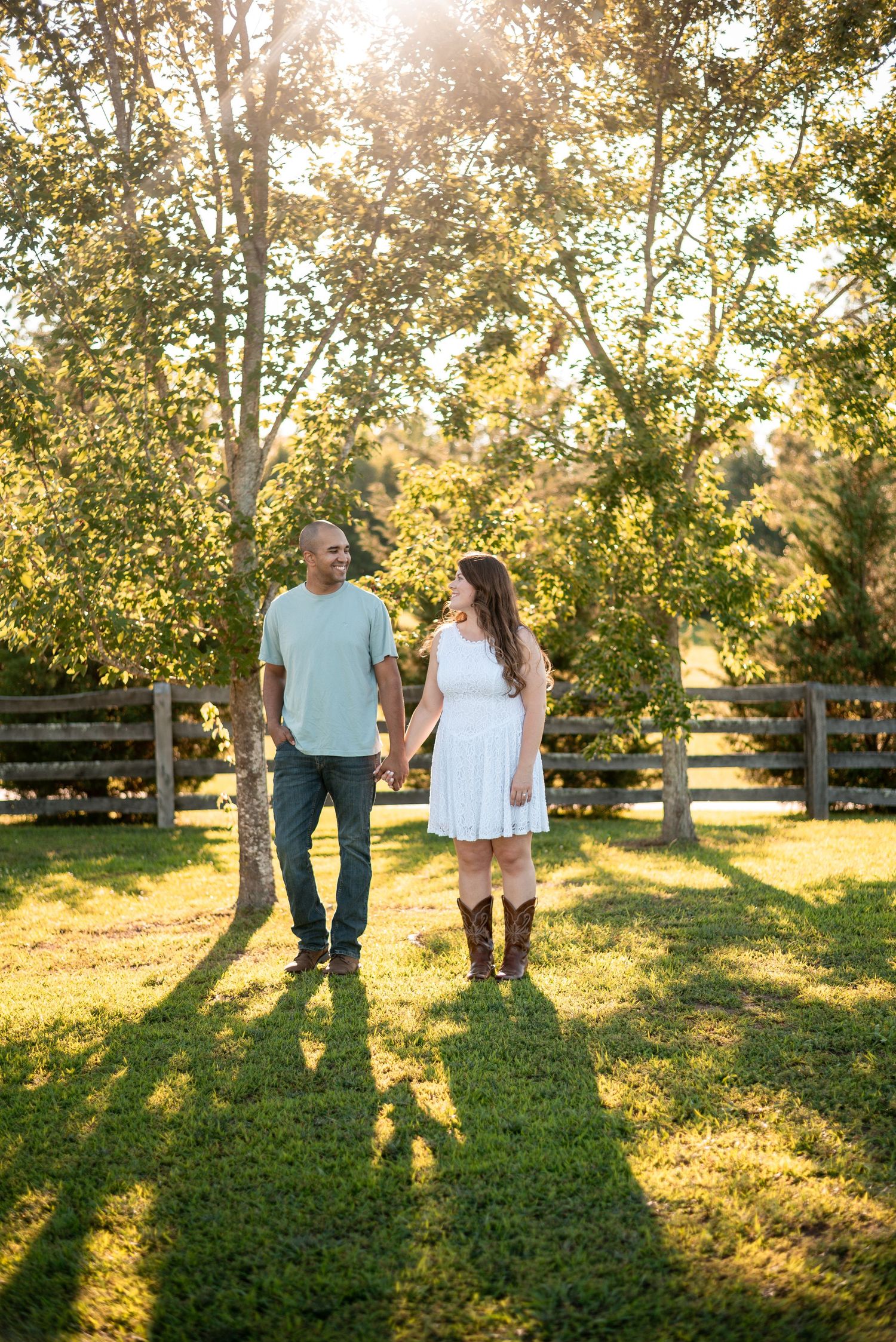 Couple holding hands and smiling at each other in a sunny park with wooden fence and trees in background.