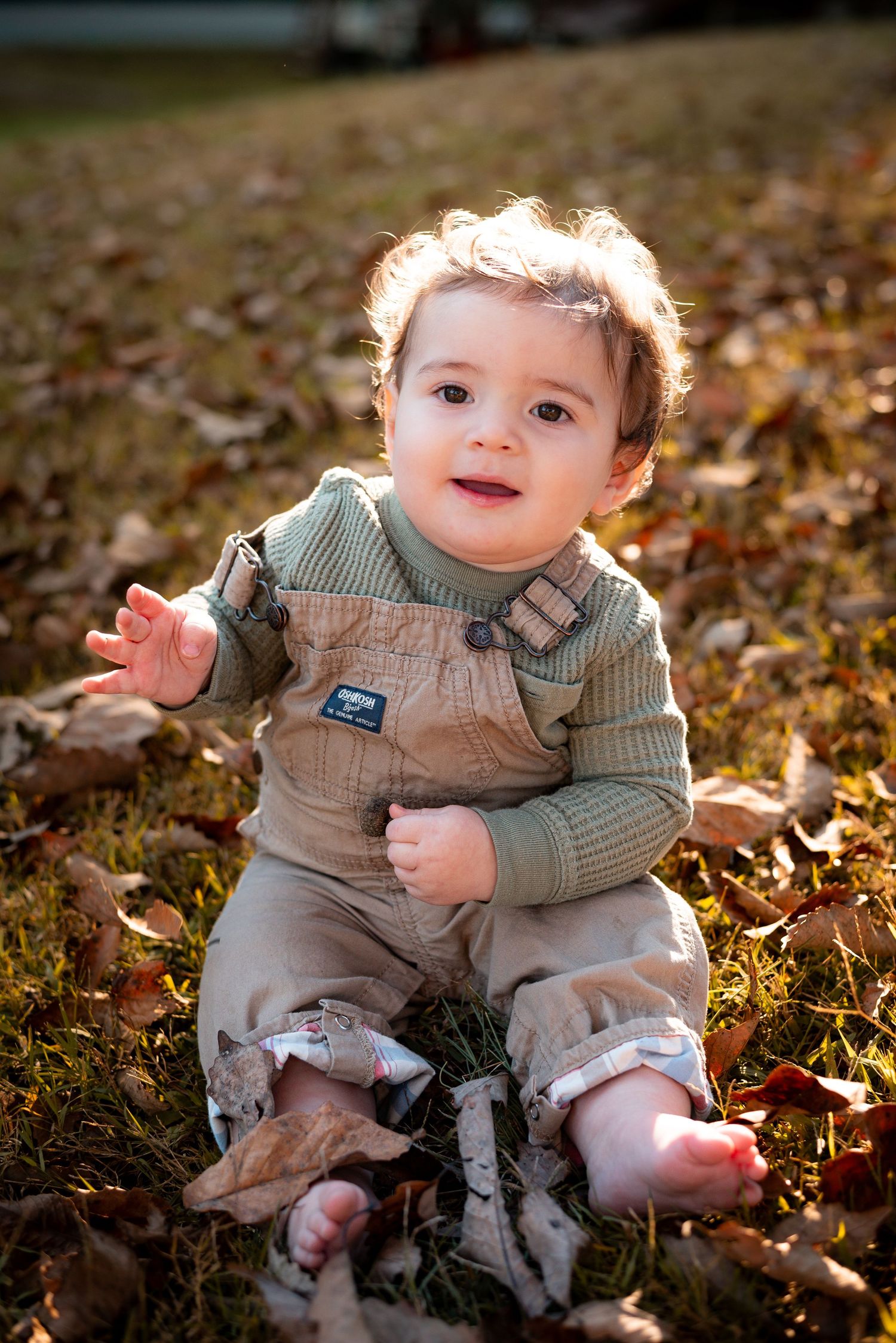 Baby in knit sweater and overalls sits among scattered autumn leaves.