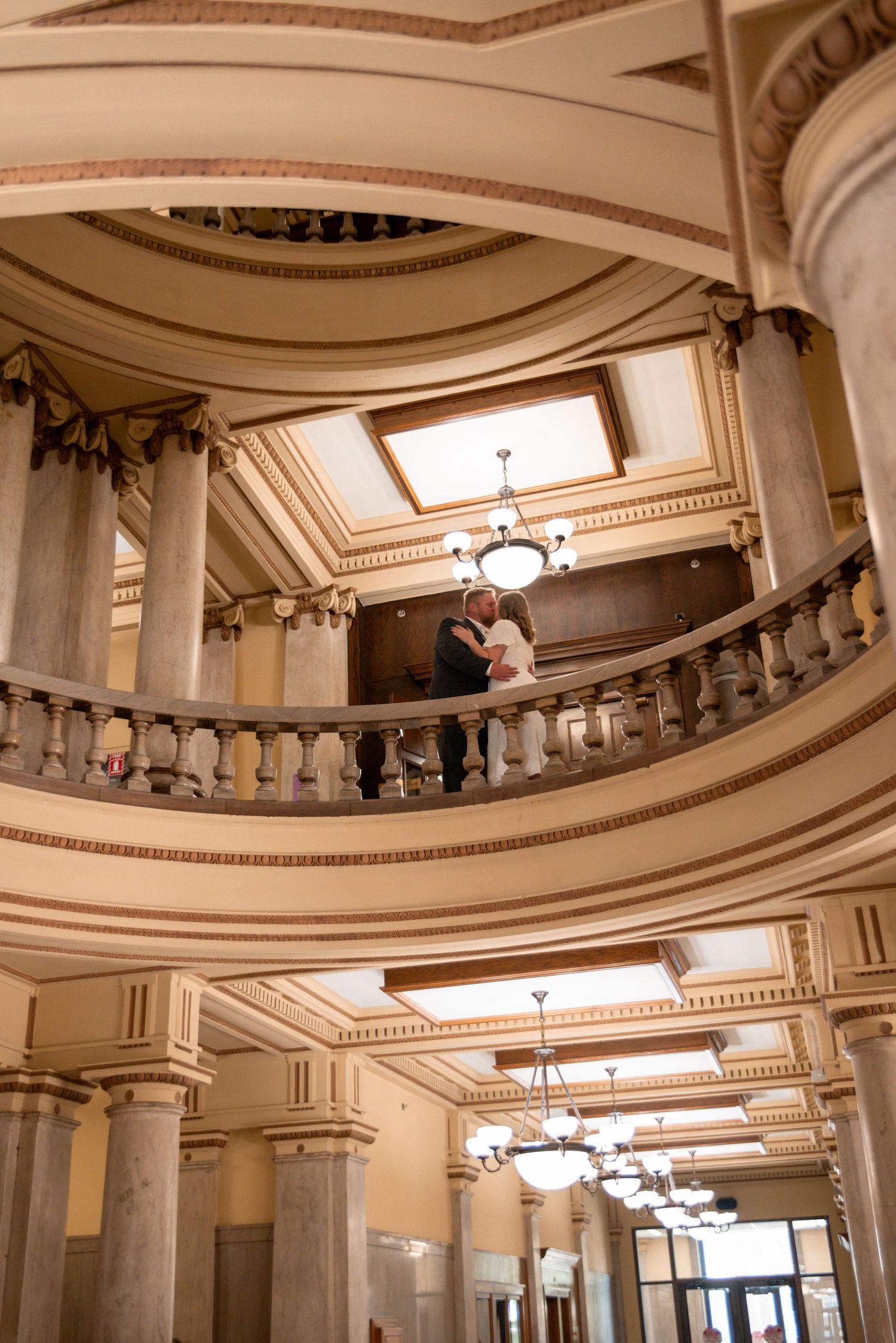Couple sharing a romantic kiss on an ornate balcony inside a grand classical building with columns and chandeliers.