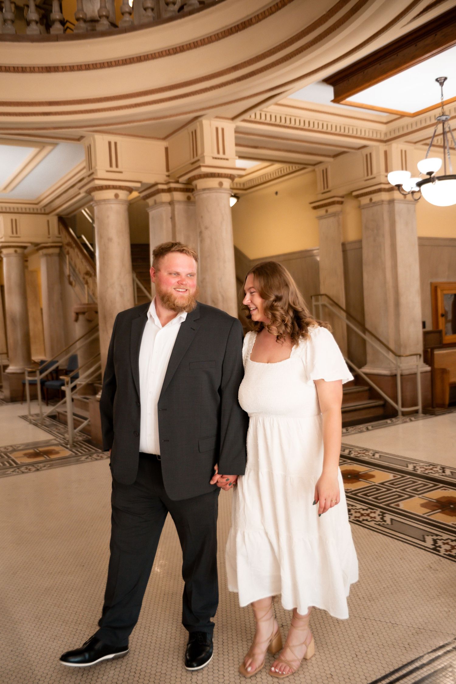 Couple holding hands smiling in elegant marble building with columns, man in black suit, woman in white dress.
