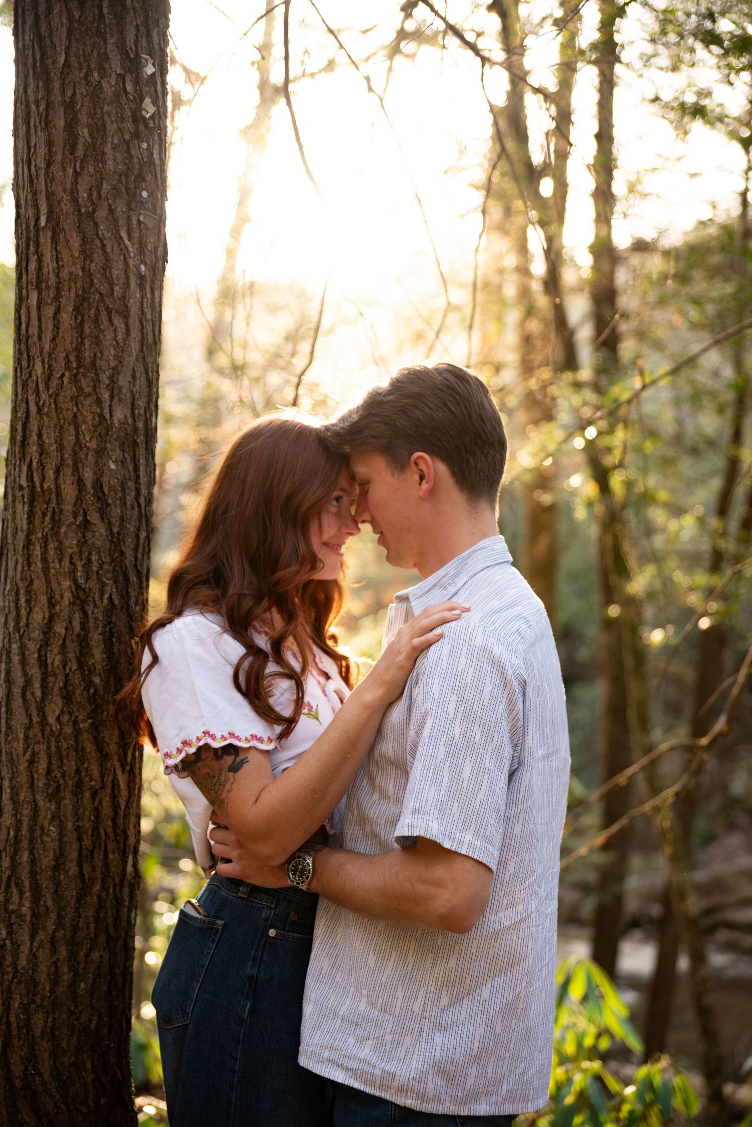 Couple sharing a romantic moment in golden sunlit forest, woman with red hair embracing man near tree.