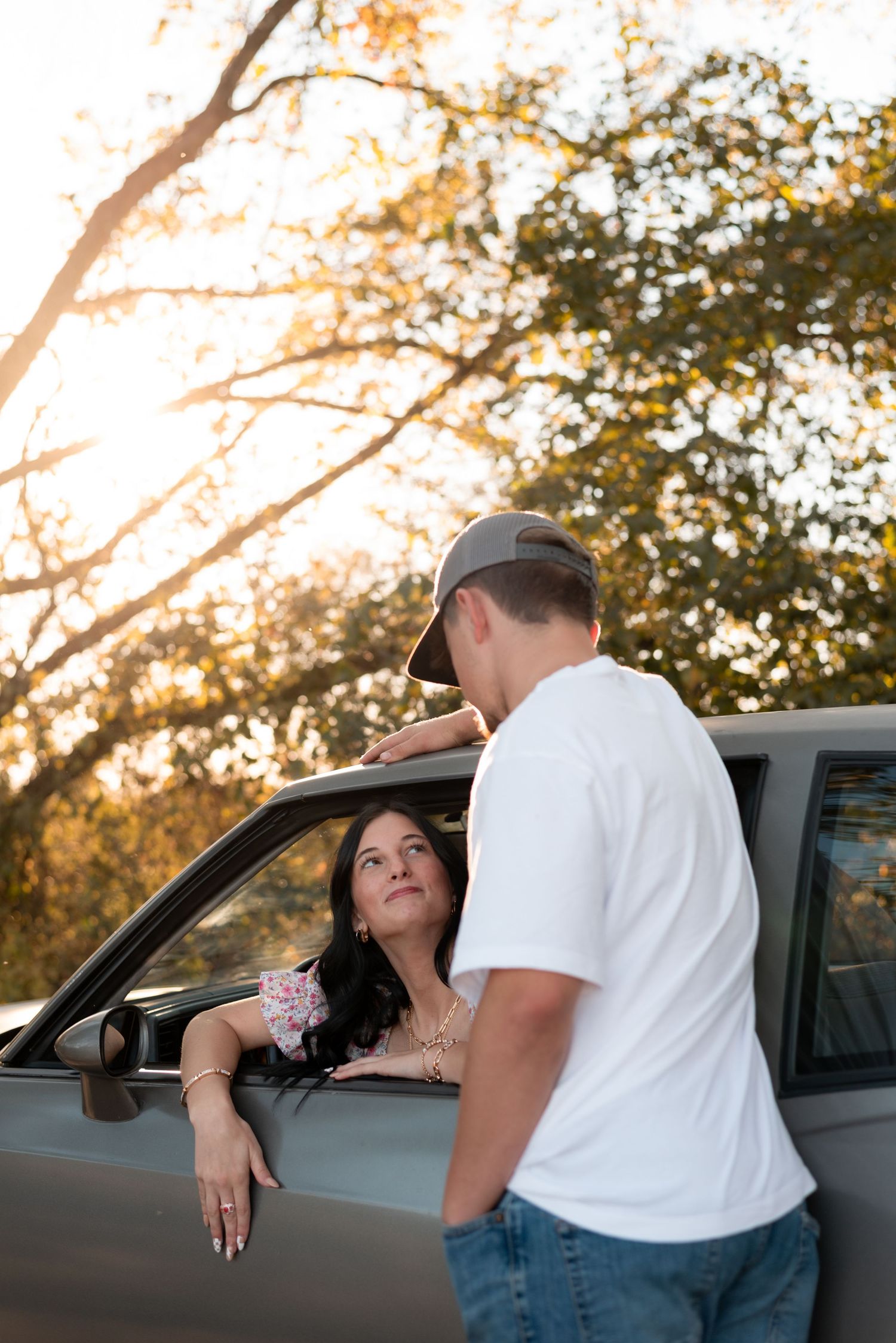 A romantic scene unfolds near a parked car during sunset with trees in the background.