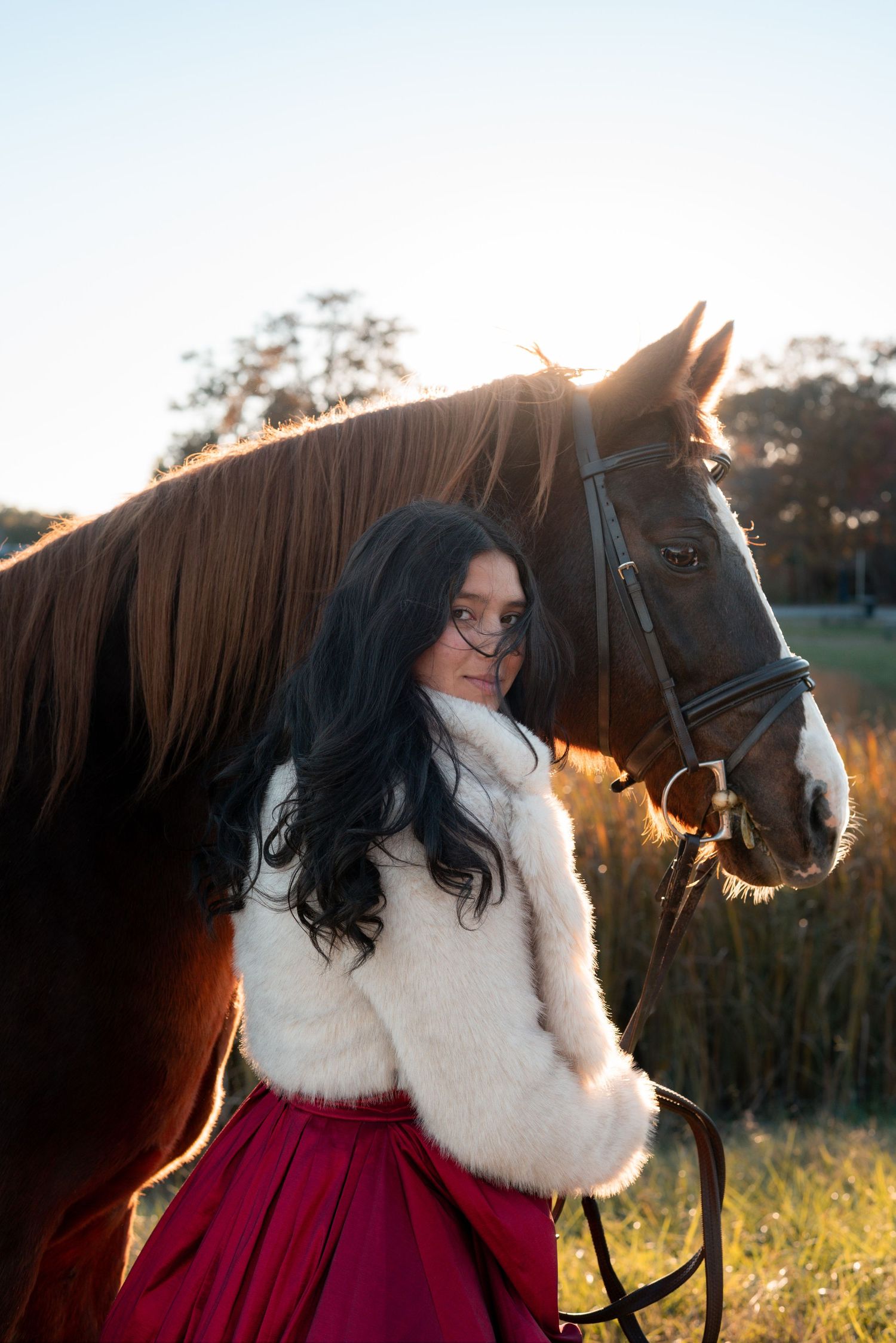 A person in a white sweater and red skirt poses with a dark horse in golden sunlight.